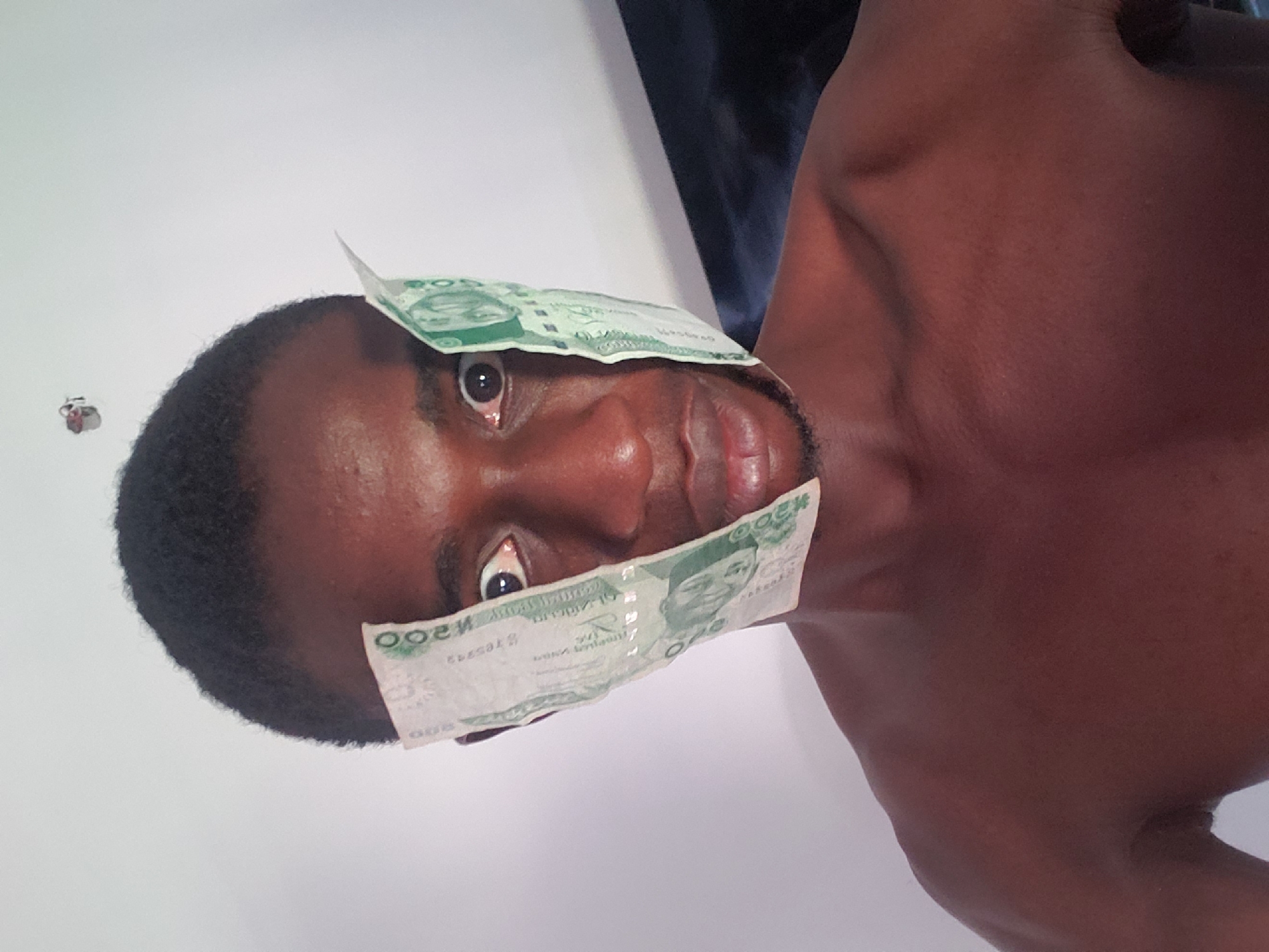 The image, oriented sideways, features a person with dark skin and short dark hair, possibly male, looking directly at the viewer. Two Nigerian Naira banknotes, likely 200 Naira notes given the visible "N200" text in reverse, cover the person's eyes. One note rests across the forehead, partially covering the upper part of the eyes, while the other is positioned across the lower face, covering the lower part of the eyes and upper cheeks. The person's right eye (viewer's left) is visible between the banknotes, appearing wide open, conveying a surprised or bewildered expression. The mouth is slightly agape. The person's bare chest and right shoulder are visible, suggesting they are shirtless or wearing attire that exposes the upper torso. The background is a plain, light-colored wall, possibly white. A darker, indistinct area is visible behind the person's shoulder, possibly furniture or clothing. The lighting is bright and even, suggesting an indoor setting during the day. The activity depicts a pose with money obscuring vision, possibly symbolizing being "blinded by money" or a similar artistic concept. The location context provided is Adonkolo, Nigeria.
