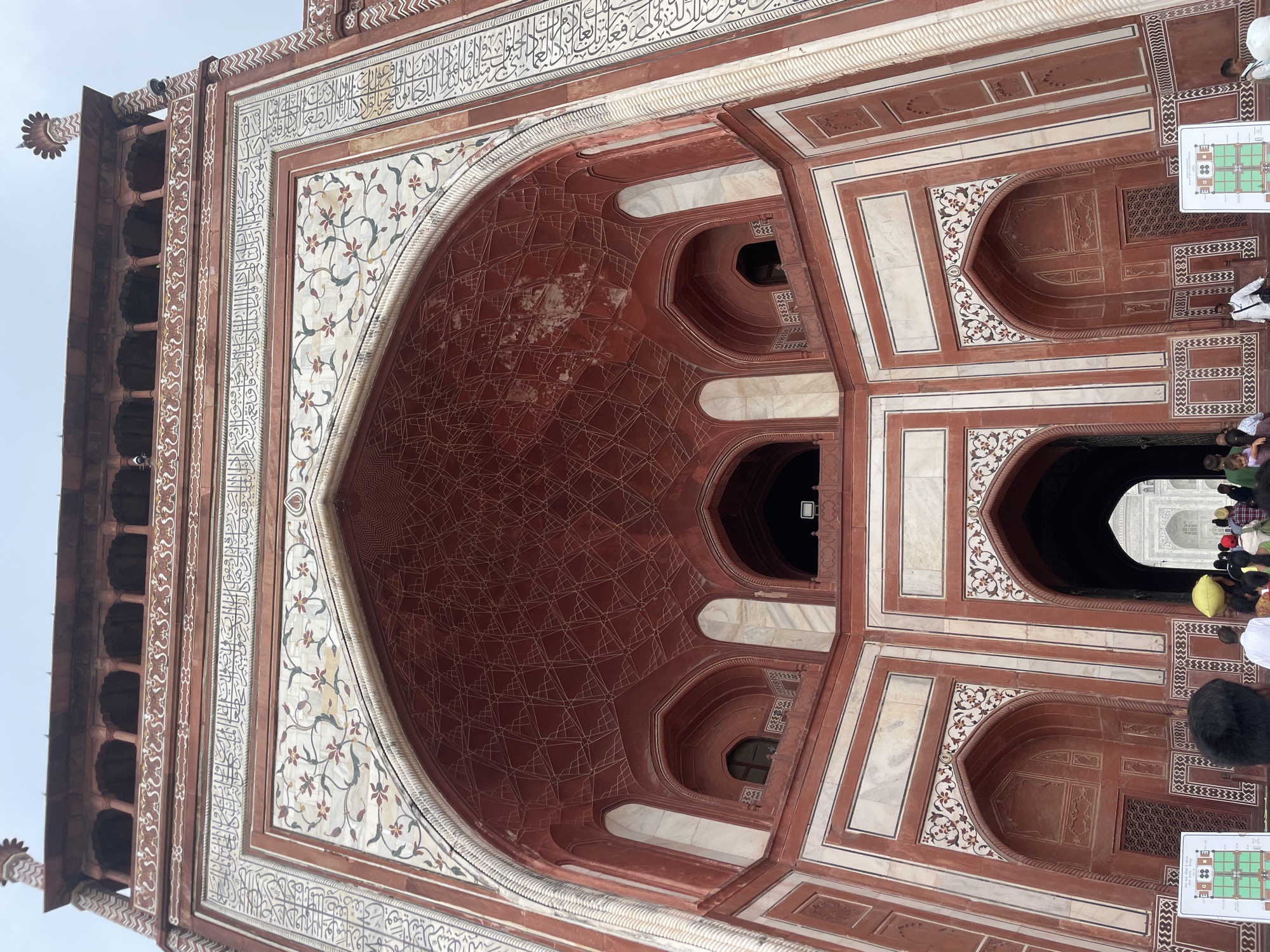 This is a low-angle, full-shot, eye-level photograph of the Darwaza-i Rauza, the main gateway to the Taj Mahal in Agra, India. The imposing red sandstone structure is richly decorated with white marble inlays, featuring floral motifs and intricate calligraphy. The grand archway of the gateway dominates the foreground, with smaller arched entrances and windows visible within its recesses. A crowd of people, some carrying yellow head coverings, are gathered in front of the gateway, preparing to enter or having just exited. The sky above is overcast, casting a soft light on the scene. The detailed carvings and symmetrical design of the gateway highlight the architectural grandeur of the Taj Mahal complex.