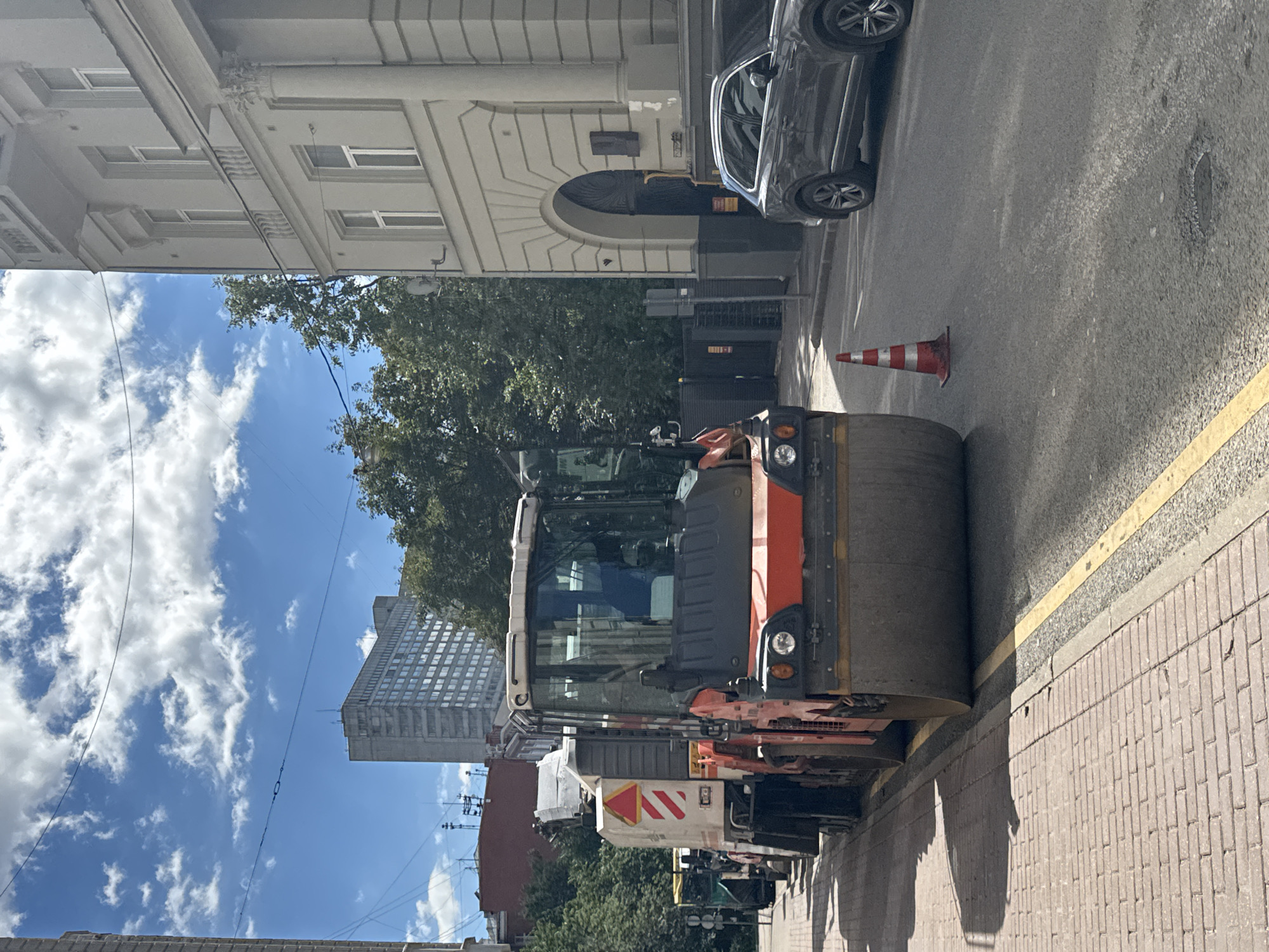 The image depicts a street scene in Moscow, Russia, on a sunny day with scattered clouds. In the foreground, a large orange and gray steamroller is parked on the right side of a paved road, its massive roller occupying a significant portion of the frame. The road has a yellow dividing line. Adjacent to the road is a brick sidewalk. In the background, a modern building with many windows stands tall, while older, multi-story buildings with classical architectural elements, including arched entrances and columns, are visible to the right. Trees line the street, and a dark-colored SUV is parked on the road. A single orange traffic cone with white stripes is placed on the asphalt. The overall impression is of ongoing roadwork or maintenance in an urban environment.