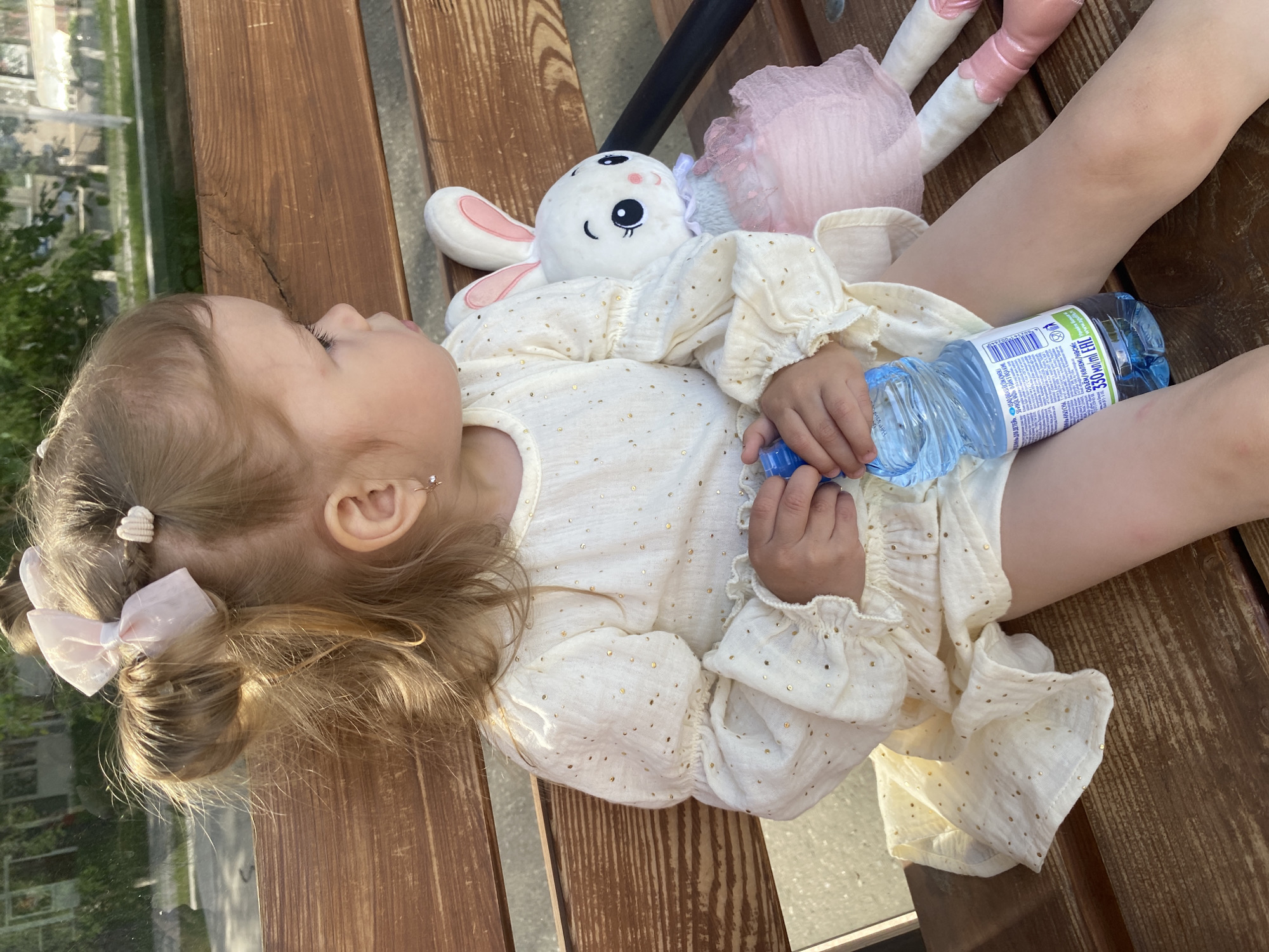 A young child lies on a wooden bench, clad in a light-colored dress adorned with gold dots. The child is clutching a plastic water bottle and a white stuffed rabbit. A pink doll also rests on the bench near the child. The location is in Chaikovsky, Russia.