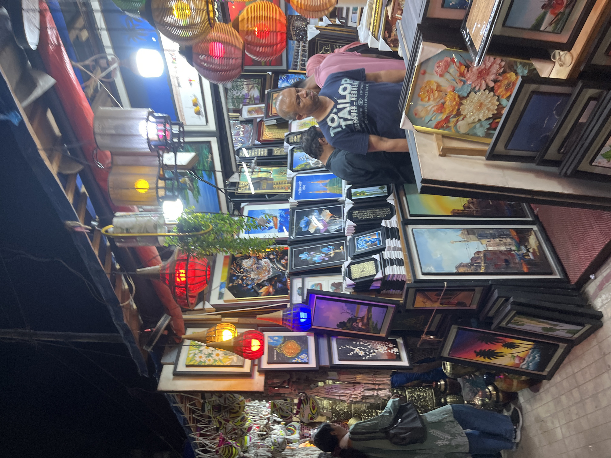 The image shows a street scene in Dhaka, Bangladesh. Framed paintings are displayed for sale. Two individuals are examining the art. Hanging lanterns add color to the scene. The environment is a crowded marketplace.