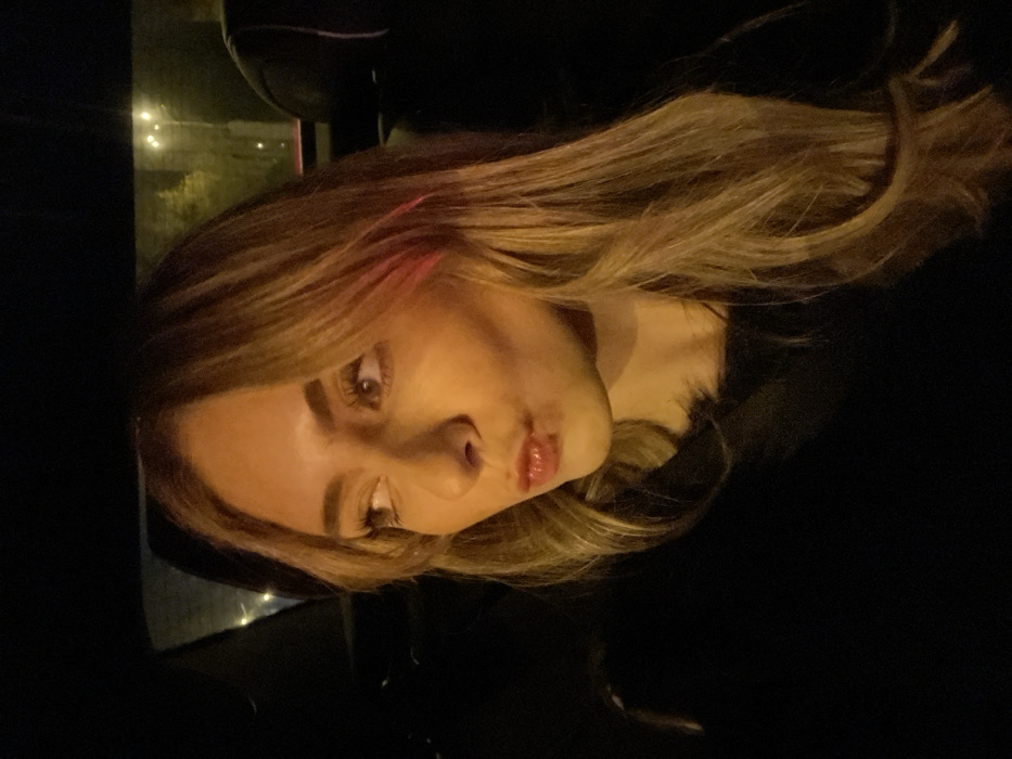 The image captures a close-up portrait of a young woman inside what appears to be a car at night. Her face is turned slightly to the left, and her head is tilted sideways within the frame. She has long, wavy blonde and light brown hair, which flows around her face and extends across the right side of the image.
The woman has distinct facial features, including defined eyebrows, open eyes that look towards the viewer, and lips slightly parted in a pouting expression, enhanced by lip gloss. A subtle reddish hue or reflection is visible on a strand of her hair near her temple.
The setting is dark, characteristic of nighttime. Through a window in the background, blurred artificial lights are visible, suggesting an urban environment. A dark object, possibly a car seat or headrest, can be seen in the upper background. The lighting is dim but casts a warm, yellowish glow on her skin and hair. There is no visible text. The activity depicted is a posed portrait or selfie taken in a vehicle in Madrid, Spain.