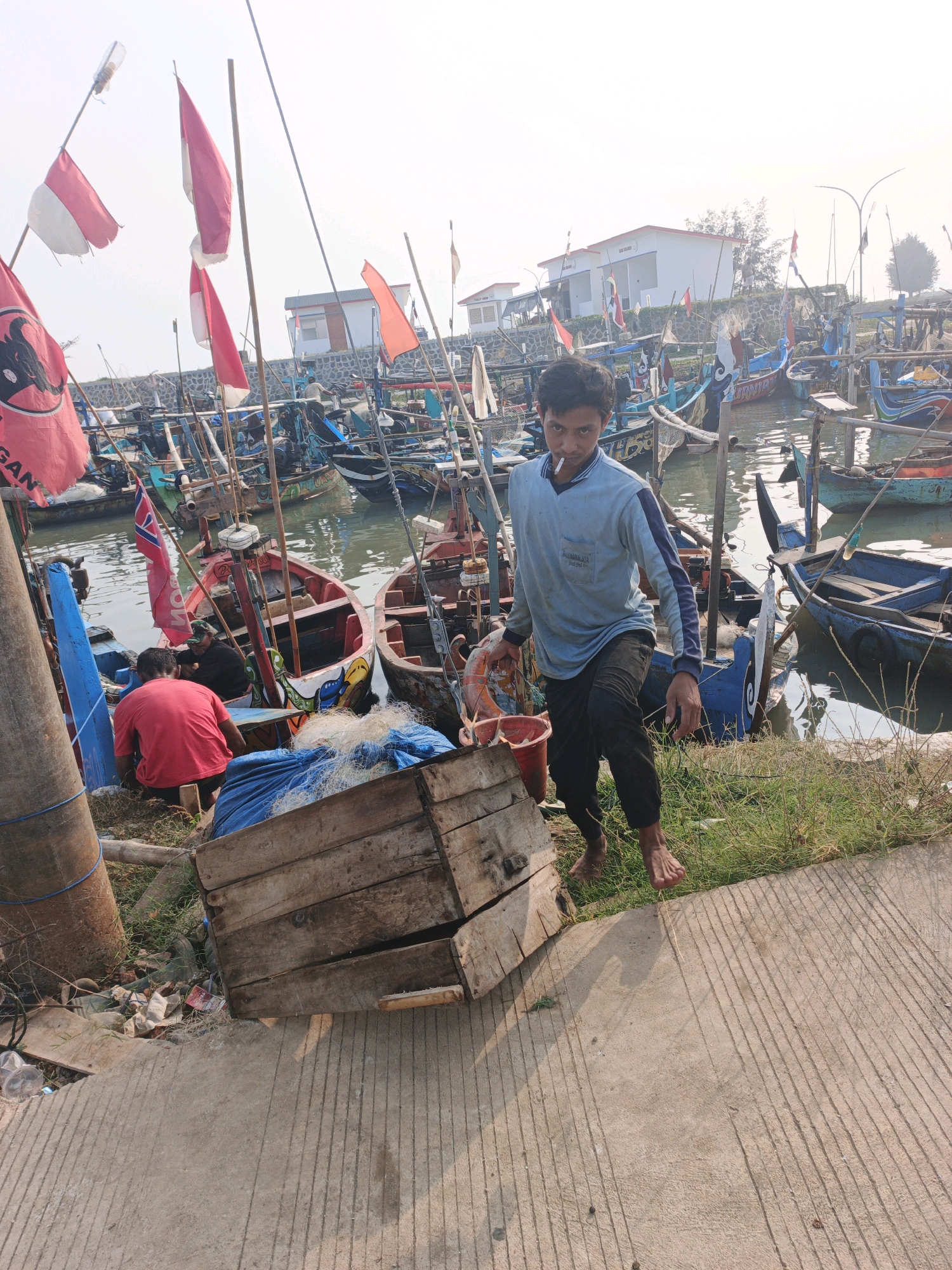 A man is walking down steps in a harbor filled with fishing boats. He is holding a cigarette in his mouth and a red bucket in his left hand. He is wearing a light blue long-sleeved shirt and dark pants. The harbor is lined with a stone wall and beyond that are two white buildings with red roofs. There are several red and white flags on poles at the harbor. A wooden crate and fishing nets are on the ground near the steps.