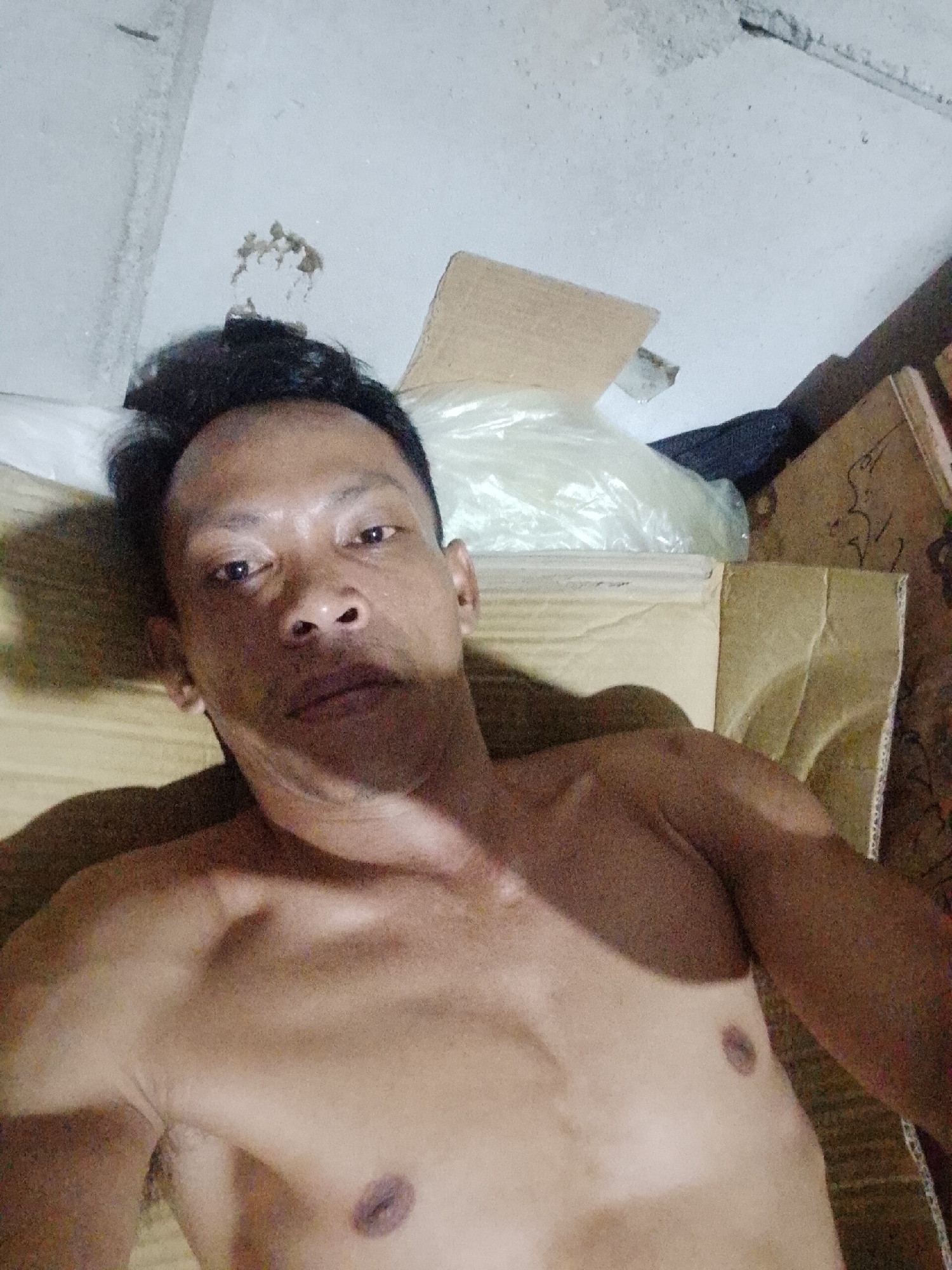 A shirtless Asian man is lying down on cardboard in an indoor space. The ceiling is visible, appearing to be bare concrete with some damage. The environment seems cluttered with items covered in plastic bags and cardboard boxes. The man is the primary subject, with his torso and head being prominent features. There's no immediate indication of a specific action or interaction beyond the act of lying down. The location is Bukit Ketok, Indonesia.