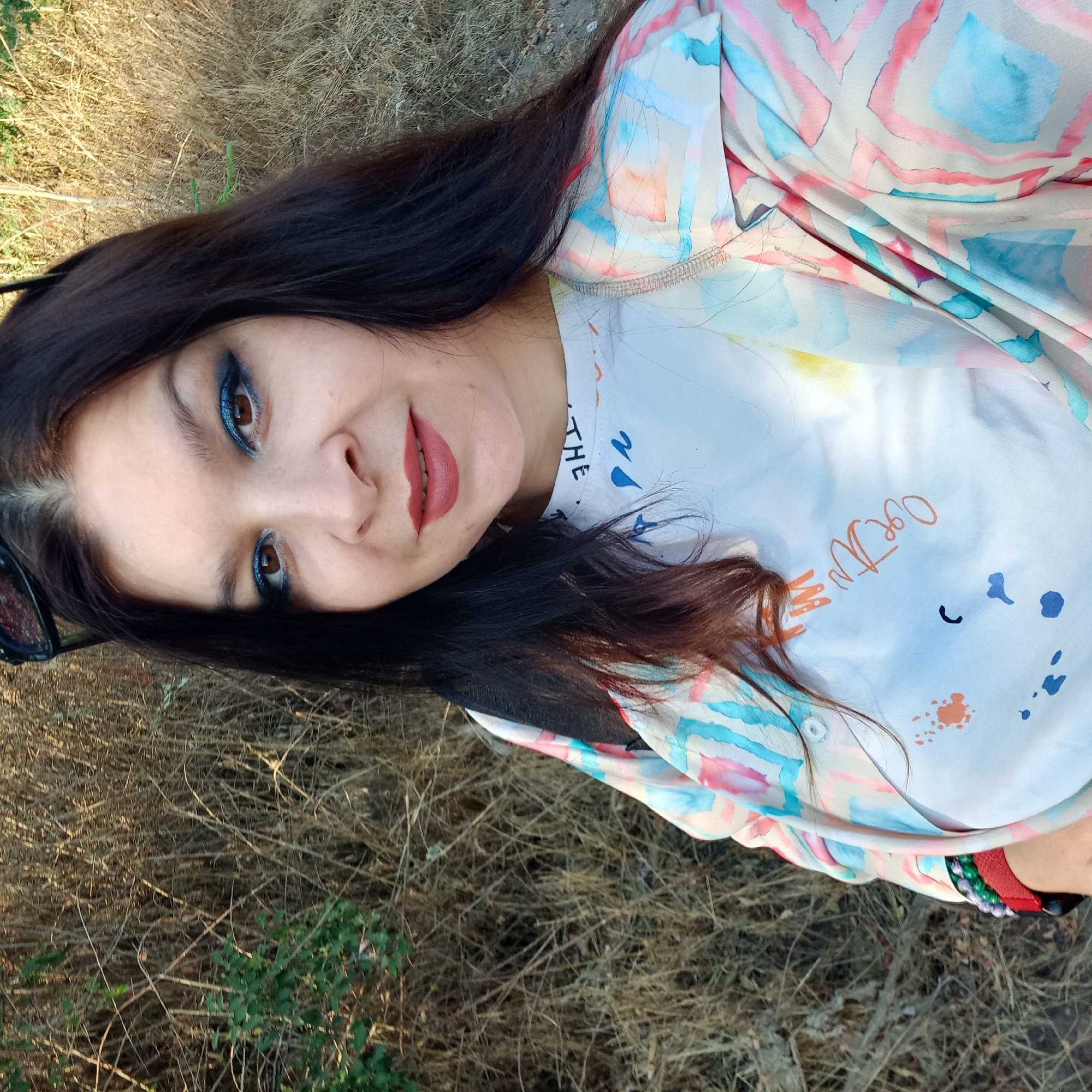 A woman with dark hair and bright blue eyeshadow smiles for the camera, her face framed by dry, tall grass. She wears a white t-shirt with abstract designs and a light-colored, patterned jacket. Sunglasses are perched on top of her head. The background consists of a dense patch of dried vegetation, suggesting an outdoor, possibly rural or natural setting. No specific location in Klyntsi, Ukraine, can be determined from the image.