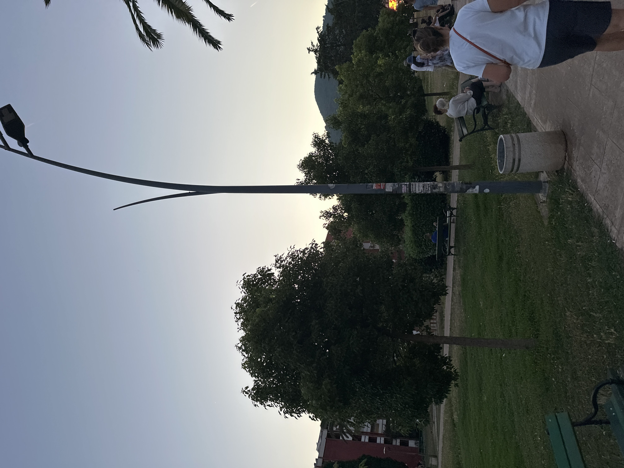 The image depicts a park scene in Budva, Montenegro, at dusk. A tall, slender lamppost with a single light fixture stands prominently in the foreground, casting a faint glow. To the left, a large, leafy tree dominates the mid-ground, with a smaller tree planted in front of it. A residential building with red accents is visible behind the trees. To the right, several people are present, some walking along a paved pathway, and others sitting on park benches. A woman wearing a white t-shirt and dark shorts is walking away from the camera, her back to the viewer. In the background, a lush green hillside with trees can be seen, bathed in the soft light of the setting sun. The overall atmosphere is peaceful and serene, suggesting a leisurely evening in the park.