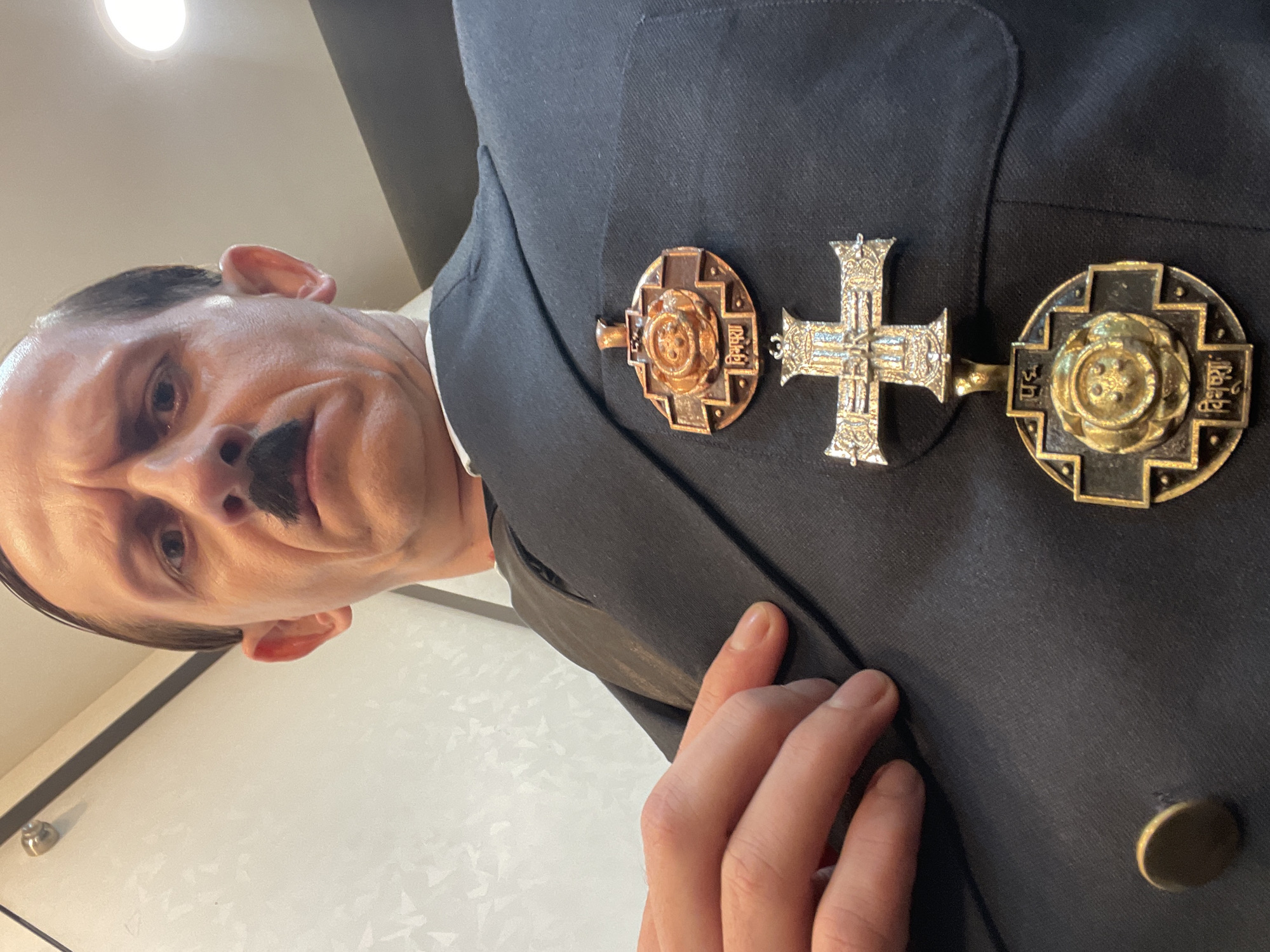 A close-up, low-angle, self-portrait shot captures a person dressed in a dark suit with medals affixed to the lapel. The individual has a prominent mustache and is looking directly at the camera with a grimacing expression. The background appears to be an indoor setting with a patterned wall and a ceiling light visible. The medals are intricate, with one being a cross shape and the other a circular design with what looks like text in an Indian script. The person's left hand is visible, resting on the suit jacket near the medals. The overall impression is that of a theatrical or costume portrayal. There is no indication of Mumbai, India, in the image.