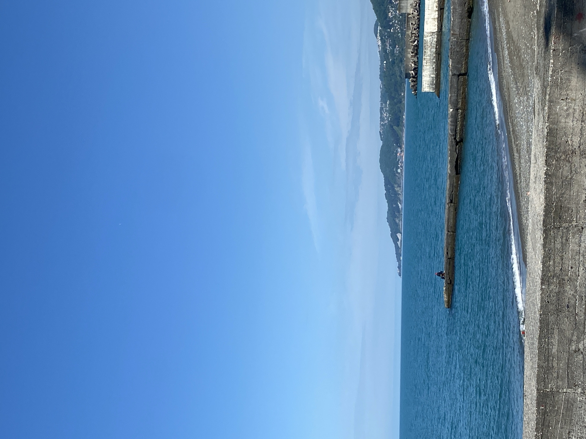 The image depicts a view of the sea, likely the Black Sea, with a concrete breakwater structure extending into the water. The structure is made of large, rectangular blocks and appears to be partially submerged, with waves gently breaking against its edge. Adjacent to the breakwater is a darker, sandy beach. The sea stretches out to the horizon, transitioning from a darker blue near the breakwater to a lighter, almost turquoise hue further out. In the distance, the coastline is visible, with buildings and vegetation dotting the landscape. The sky is a clear, bright blue, occupying a significant portion of the frame. On the breakwater, a figure can be seen. Given the coastal features and the style of the breakwater, it is plausible that this scene could be located in or near Sochi, Russia, which is a coastal city along the Black Sea.