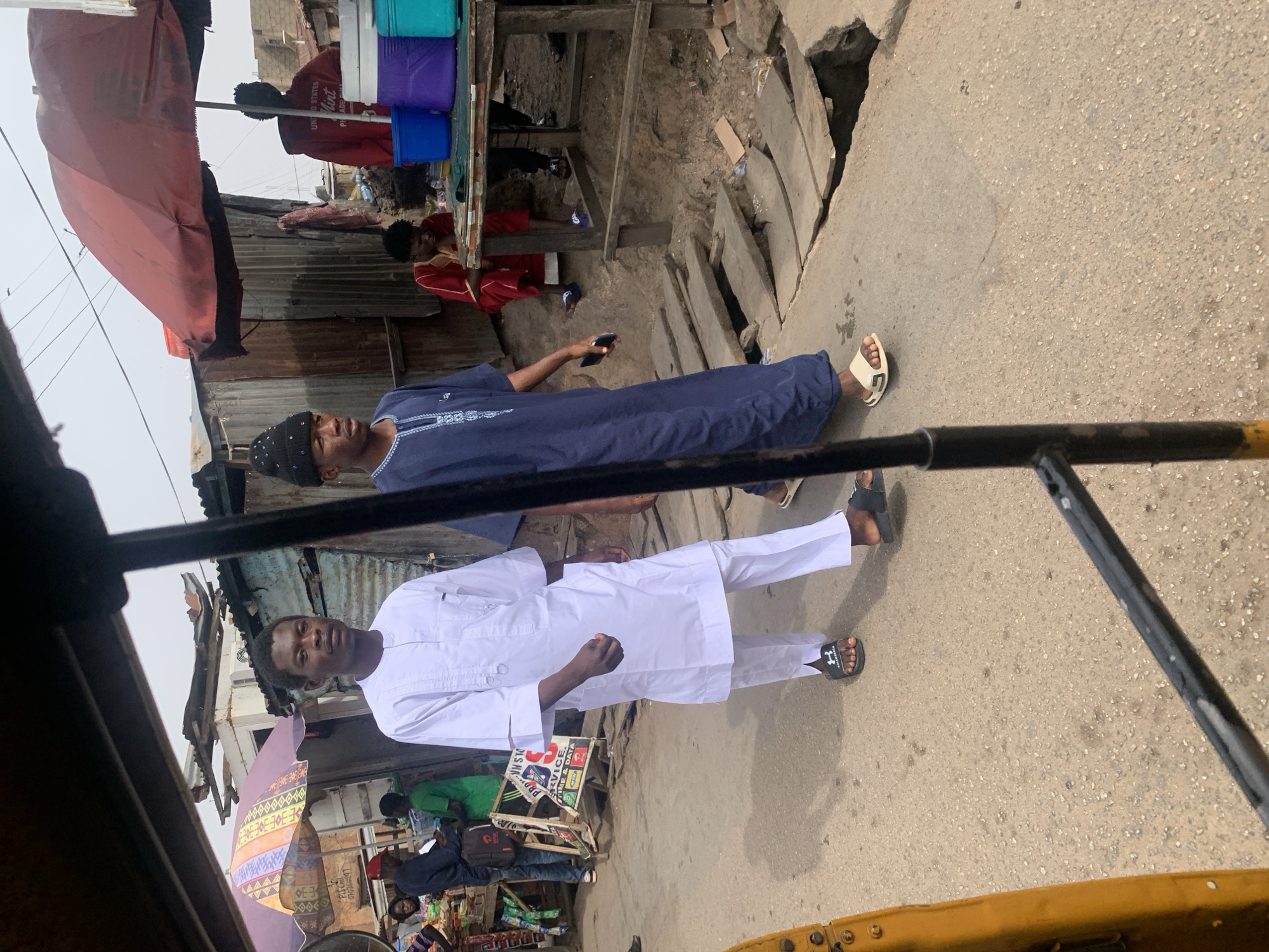 The image captures a vibrant street scene, likely in Jos, Nigeria, under bright daylight conditions. The perspective suggests the photo was taken from within a keke (auto-rickshaw), with a black metal frame visible across the foreground.
In the center, two men stand on a concrete/asphalt road. Closer to the viewer, a man in a white traditional outfit (kaftan/agbada style) with matching trousers and black Under Armour sandals looks directly towards the camera. Beside him, slightly further back, is another man wearing a dark blue traditional tunic with light embroidery, a black beanie, and white sandals. He is holding a mobile phone.
The background reveals a bustling market or commercial area. Structures are primarily constructed from corrugated metal sheets and wood, forming makeshift stalls. Several other individuals are visible, including one person in a red shirt with the text "UNIO STATES Mint PHADE" standing behind cool boxes (light blue, purple, dark blue). Another person in a red garment is seen further back, and others are partially obscured by stalls and colorful umbrellas, including one red and one patterned umbrella. The ground transitions from a paved road to rougher terrain and an open drainage ditch to the right.
Visible text fragments on various signs include "E3", "PLS CHIL", "07036297", "PRO OLS NA", and "RVICE. TIME & DATA," likely indicating services like mobile phone data. The scene depicts everyday activity in a busy street environment.