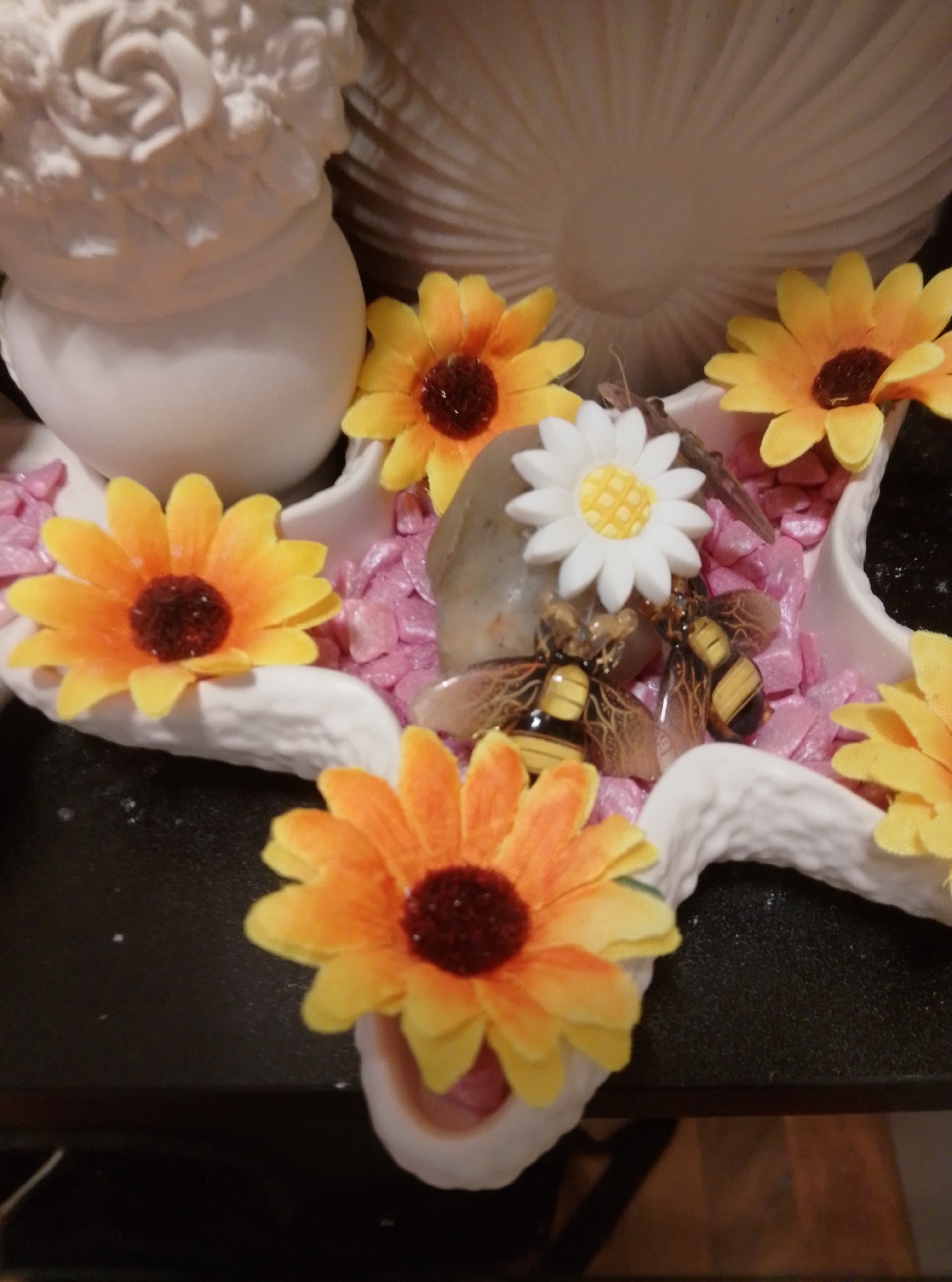 This image depicts a decorative arrangement on a dark surface, possibly a table, in a domestic setting. The arrangement features several artificial sunflowers with orange petals and dark brown centers, interspersed with small, pink, pebble-like decorative stones. Two small, decorative bee figurines, with yellow and black striped bodies and transparent wings, are placed among the flowers and stones. A single white daisy figurine with a yellow center is also present. In the background, a large, textured, off-white object resembling a decorative shell or sculpture is visible, along with a white, somewhat abstract, sculpted object that appears to be a base or part of a larger decorative piece, possibly depicting a bust or floral elements. The scene is brightly lit, highlighting the colors and textures of the artificial flowers and figurines. No individuals are visible in the image, and there is no indication of the specific location within the city of Wedel, Germany.