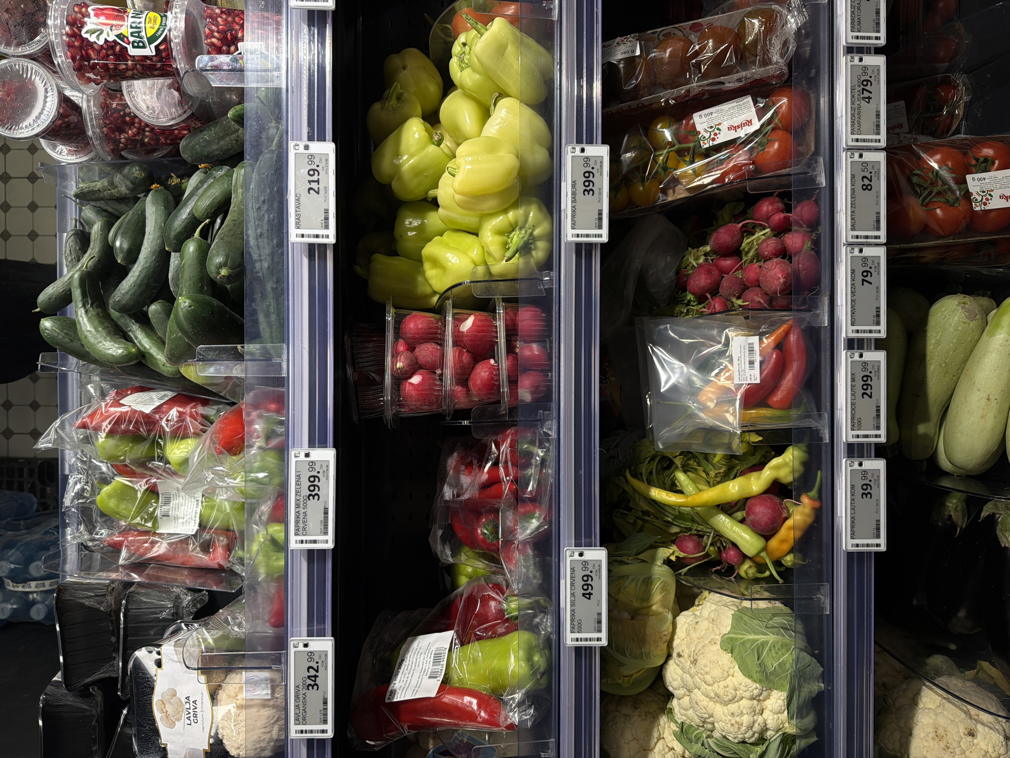 The image depicts a produce section in a supermarket, likely located in Vracar Urban Municipality, Serbia, based on the Serbian language price tags. The shelves are stocked with various fruits and vegetables, including pomegranates, cucumbers, green and red peppers (labeled as "Paprika"), radishes, tomatoes (labeled as "Paradajz"), zucchini, hot peppers, and cauliflower. The items are arranged in clear plastic containers and bags, with price tags attached to the shelf dividers. Specific items labeled include "Krastavac" (cucumber), "Paprika babura" (bell pepper), "Rotkvice veza kom" (radish bunch), and "Lavlja griva organska" (organic Lion's Mane mushroom).