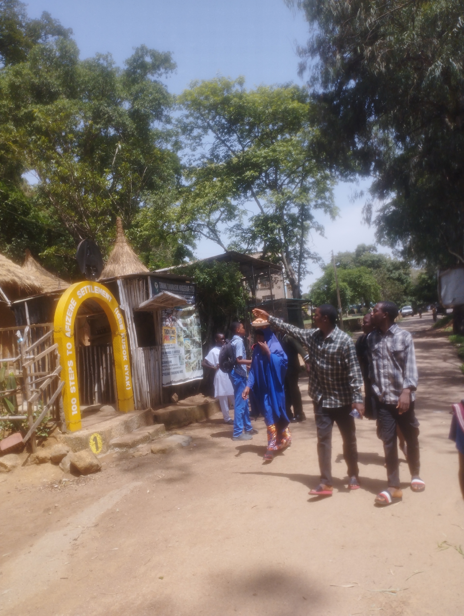 The image shows a group of people in an outdoor setting, likely in Jos, Nigeria. The scene appears to be a tourist attraction or cultural site. In the foreground, a yellow archway with text indicating "100 STEPS TO AFIZERE SETTLEMENT" and "(IGBE KUSOK NYAK)" suggests this is an entrance or pathway. Several individuals are walking on a dirt path.
One man in a plaid shirt is pointing towards something off-camera, possibly explaining the site to others. Another man in a similar plaid shirt stands next to him. Behind them, a person in a blue robe and another in white traditional attire are visible. To the left, a small building with thatched roofs and wooden walls houses information, with text visible that includes "THINK TOURISM NIGERIA-TTX," "TICKETING," and "GAME RESORT."
The environment is characterized by lush green trees overhead and a bright, sunny sky, suggesting it is daytime with fair weather. The overall impression is a lively tourist destination, with people engaging with the location and its offerings. The presence of the archway, signage, and traditional architecture points towards a cultural or historical site.