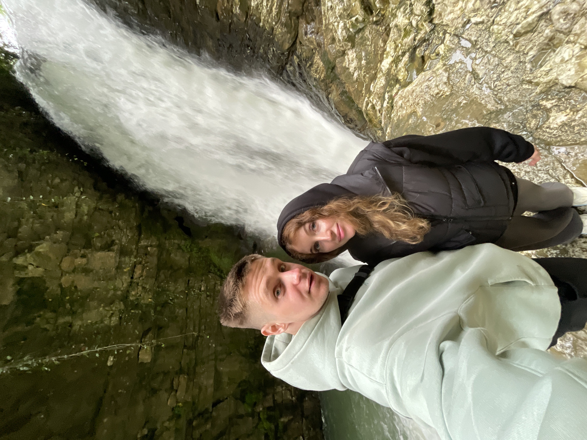 The image shows two people posing for a selfie in front of a waterfall. The man is wearing a light green hooded sweatshirt, and the woman is wearing a black hooded jacket. The waterfall is cascading down rocks covered in moss and lichen. The exact location within the city of Unknown, Unknown, is not discernible from the image.