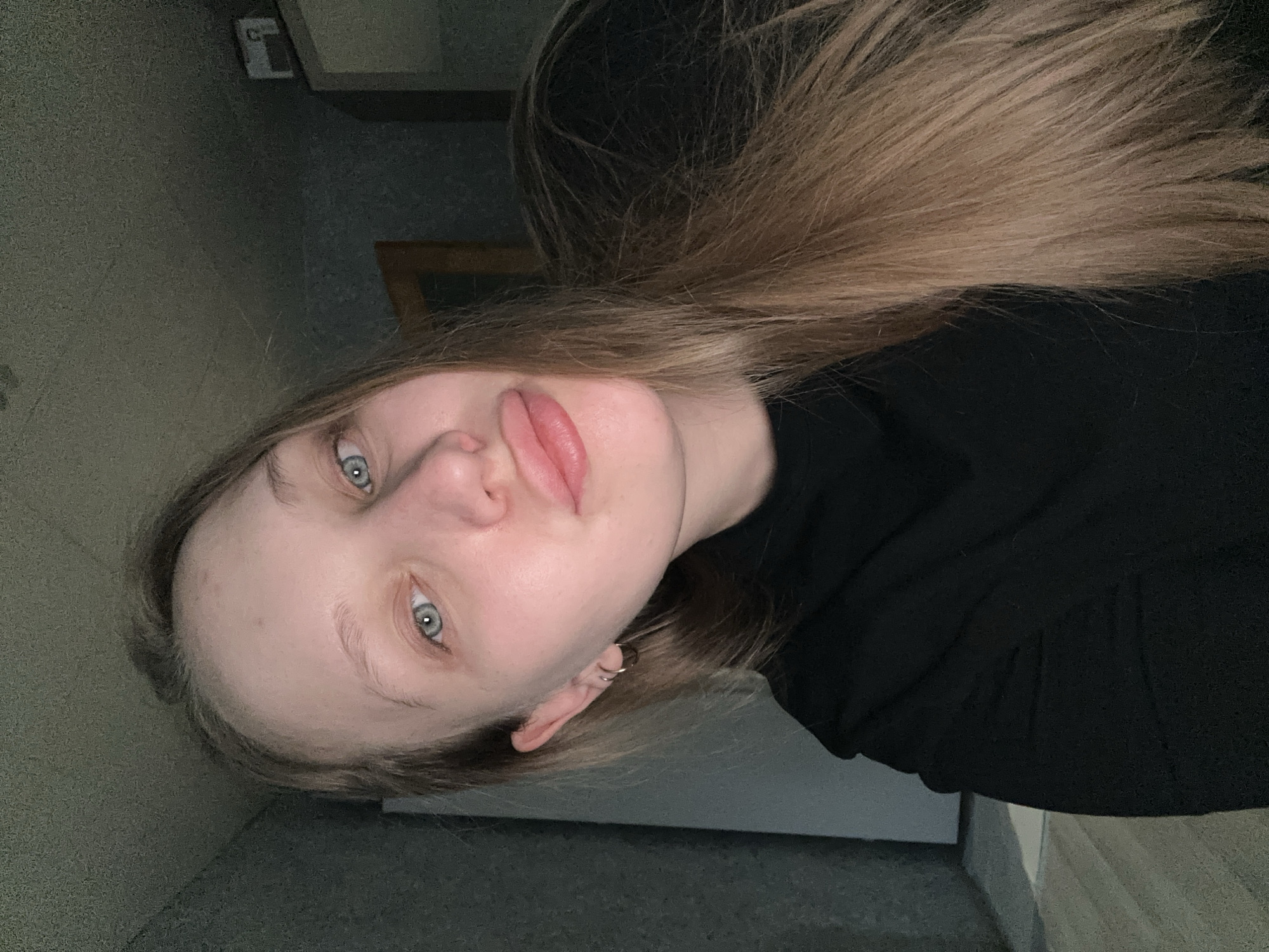 A young woman with fair skin, light-colored hair, and blue eyes is posing for a selfie indoors. She is wearing a black garment, possibly a shirt or sweater. The background suggests an interior space with textured walls and some furniture, possibly a cabinet or shelf. The location is indicated as the city of Chaikovsky, Russia, but there are no specific details visible in the image to confirm or deny this.