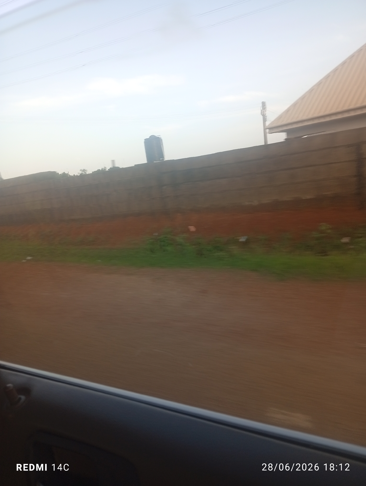 The image captures a view from a moving vehicle, looking out over a reddish-brown dirt road towards a long, grey concrete wall. Beyond the wall, sparse green vegetation and trees are visible, with a tall, slender blue building in the distance. To the right, a section of a building with a brown, sloped roof is partially visible behind the wall. Overhead, thin, dark power lines stretch across a hazy sky. The bottom of the image shows a dark grey car door frame. White text in the lower left corner reads "REDMI 14C", and in the lower right corner, "28/06/2026 18:12". The scene appears to be a roadside view, potentially on the outskirts of a city, given the presence of both urban structures and natural elements. The date and time stamp suggest the image was taken in the late afternoon.
