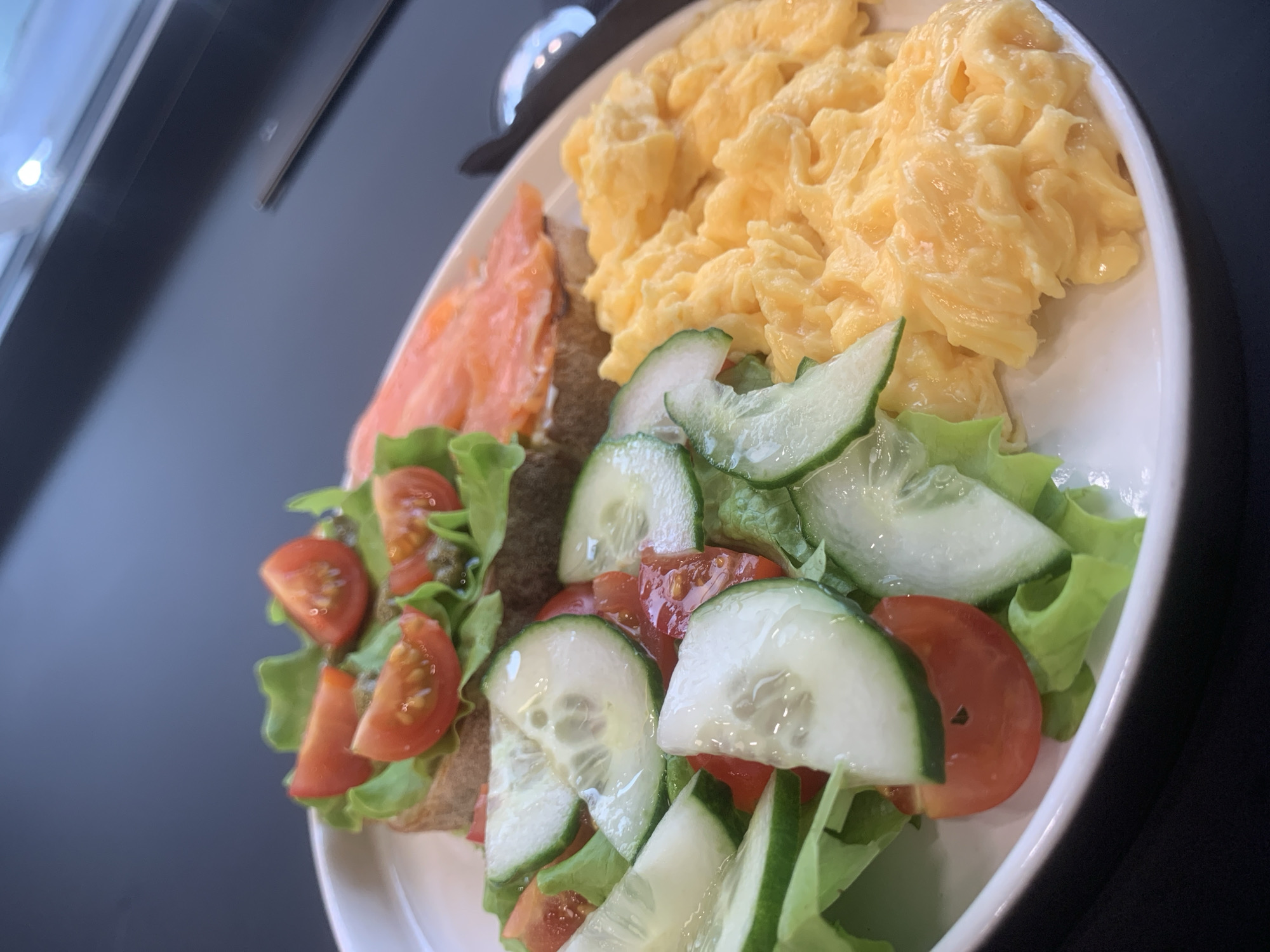 A plate of food is positioned on a dark, reflective surface. The food components include a pile of scrambled eggs, a slice of smoked salmon placed atop a piece of bread, and a salad consisting of lettuce, sliced tomatoes, and sliced cucumbers also on bread. The location is not evident from the image content. The description does not specify any people or actions.