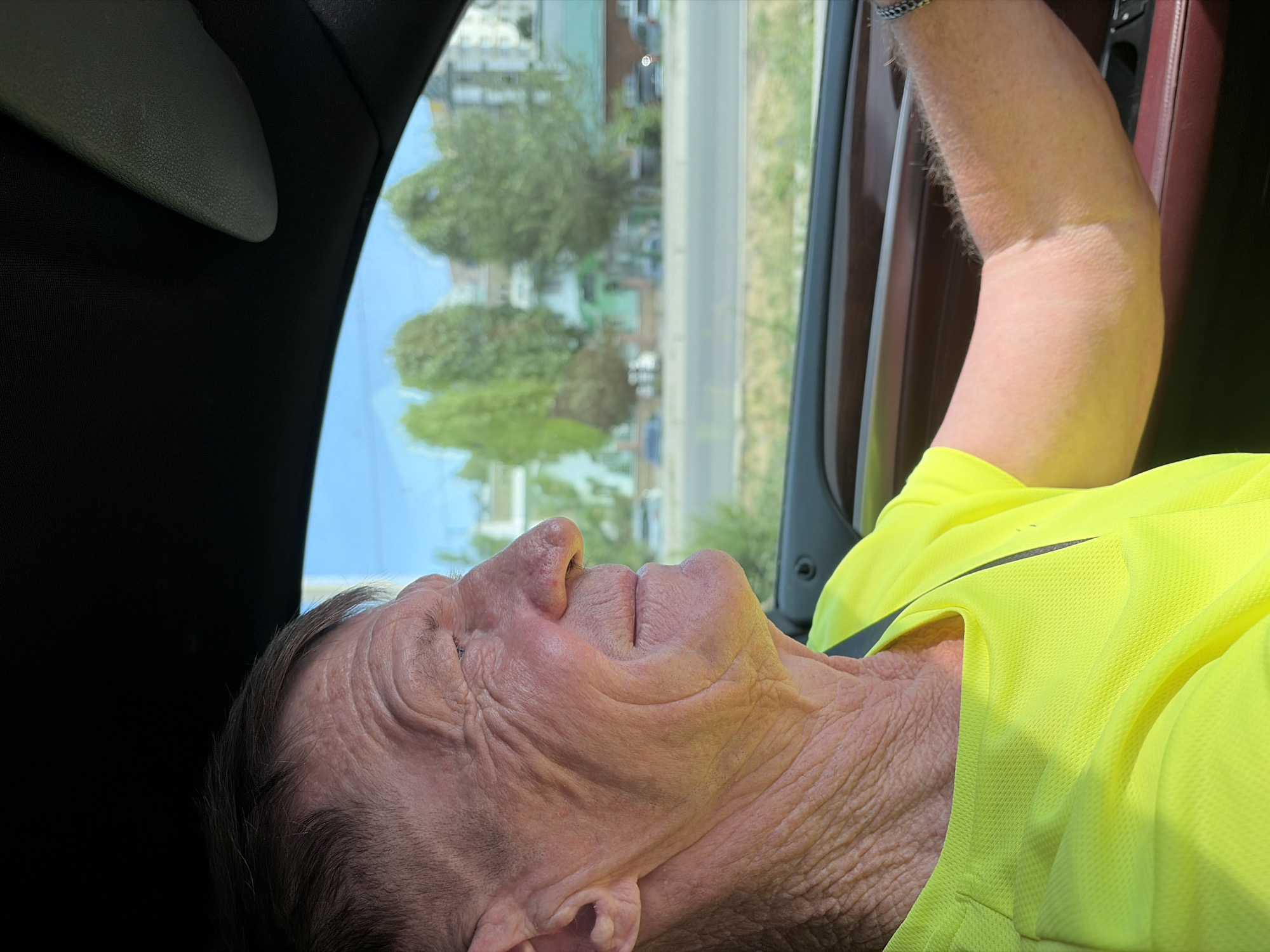 An elderly Caucasian man with wrinkled skin and short, graying hair is driving a car. He is looking to his right, with a slight smile on his face. He is wearing a bright yellow t-shirt. The interior of the car is visible, including the dashboard and the steering wheel. Outside the car, trees and buildings can be seen through the window. The sky is blue.