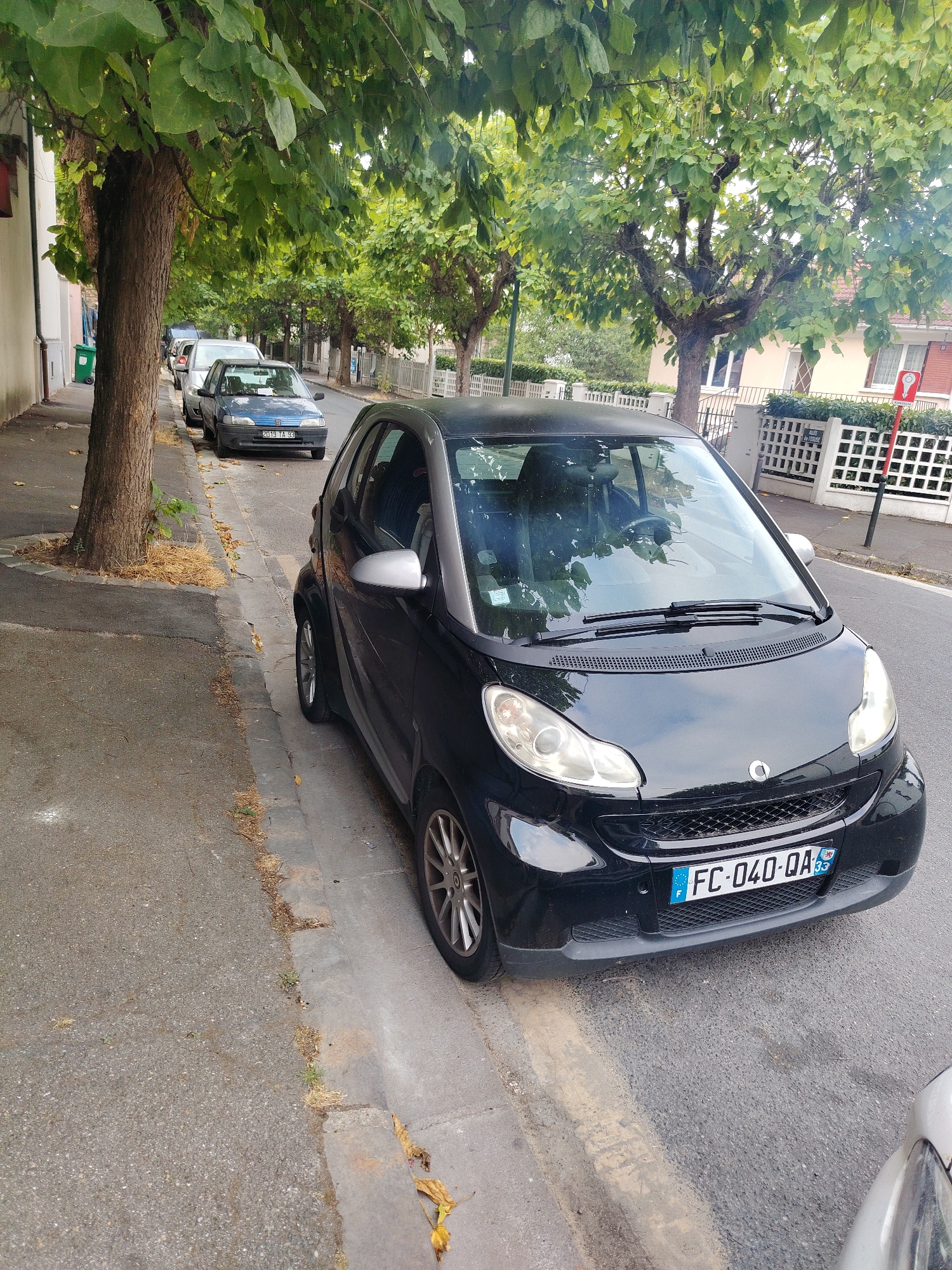 The image depicts a street scene in Clichy-sous-Bois, France. In the foreground, a black Smart car is parked along a curb. The car has a license plate reading "FC-040-QA" and a circular emblem on the front grille. Behind the Smart car, a blue Peugeot 206 is parked, with its license plate visible as "2019 TA 99". Further down the street, other vehicles are parked, and trees line the sidewalk. The environment includes residential buildings with white fences and a street sign indicating "ALLEY OF THE OLD MILL". The ground is asphalt with some dry leaves scattered on the sidewalk and road. The lighting suggests it is daytime. No people are visible in the image.