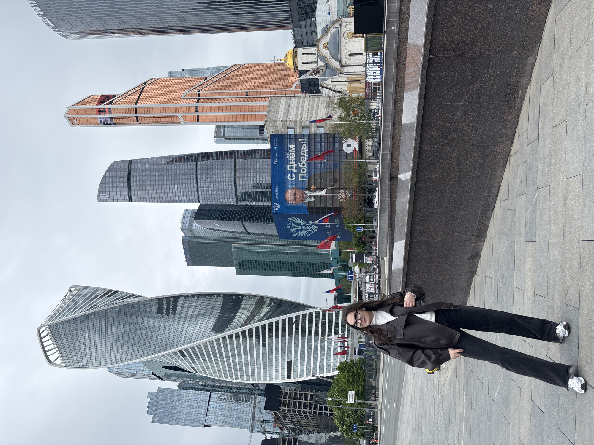 The image depicts a woman standing on a grey-paved surface, likely a pedestrian area, with the Moscow International Business Center ("Moscow City") serving as the backdrop. She is wearing a brown jacket, a white top, black pants, and sneakers.
Behind her, multiple skyscrapers are visible, including the Evolution Tower, which is known for its twisting shape. A poster with Russian text and an image of a man is situated behind a church with a golden dome. Numerous flags are planted near the poster. The weather appears overcast.