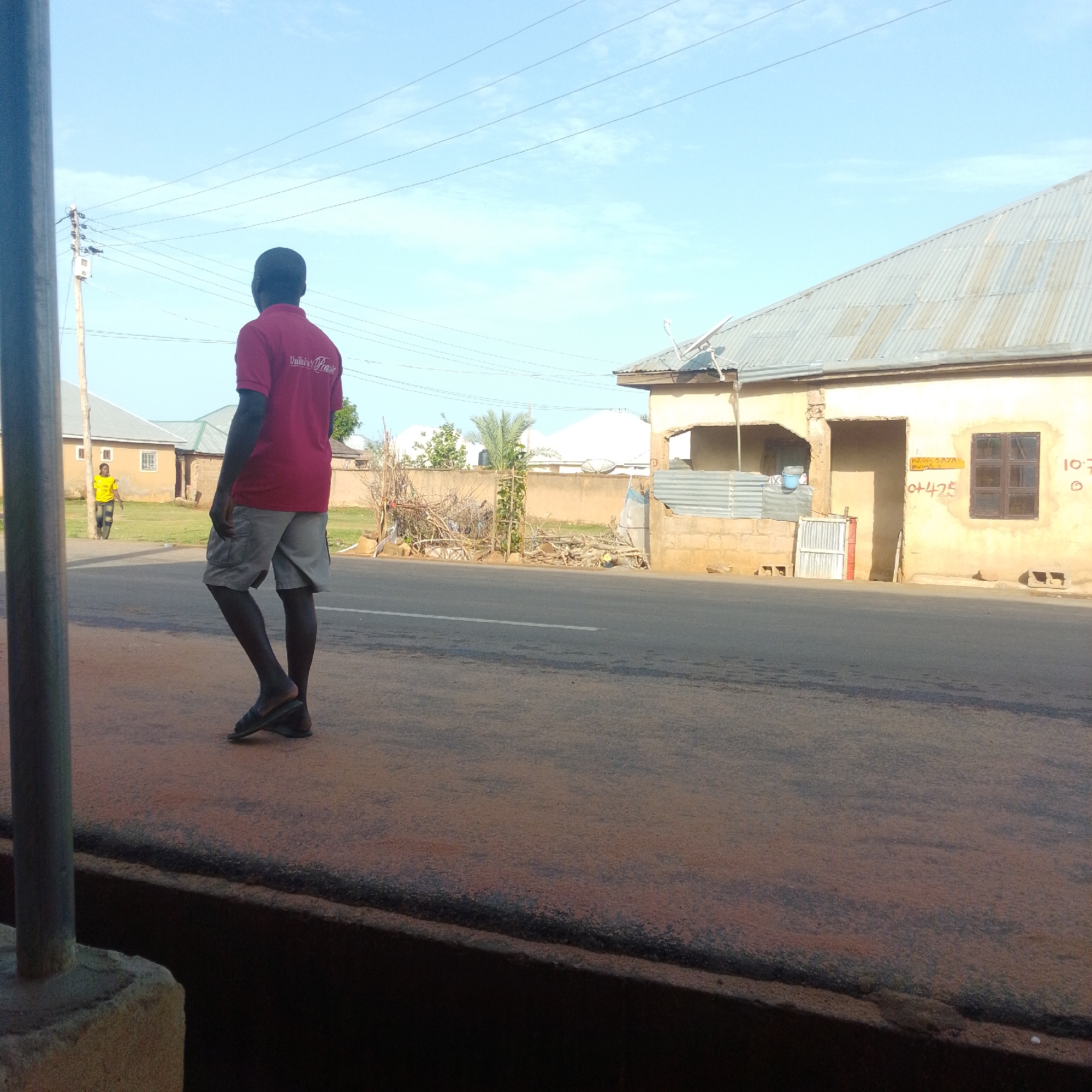 An over-the-shoulder view of a person wearing a red polo shirt with text that appears to say "Unity Praise" and grey cargo shorts, standing on a reddish-brown paved surface. The person is facing away from the camera, with their left foot slightly forward. To the left of the person, a short, grey pole stands on a concrete base. In the background, a road stretches horizontally across the frame, with white lane markings. On the far side of the road, a few buildings are visible. The building closest to the right has a corrugated metal roof and a pale yellow painted facade with some visible brickwork. There is a dark-brown window and some writing in red and yellow paint. To the left of this building, there is a pile of dry branches and vegetation. Further to the left, a smaller building with a grey roof is visible, and beyond that, a person wearing a yellow t-shirt is walking. The sky is clear and light blue with some thin white clouds. The scene appears to be outdoors in a warm climate, likely in the city of Tempure, Nigeria.