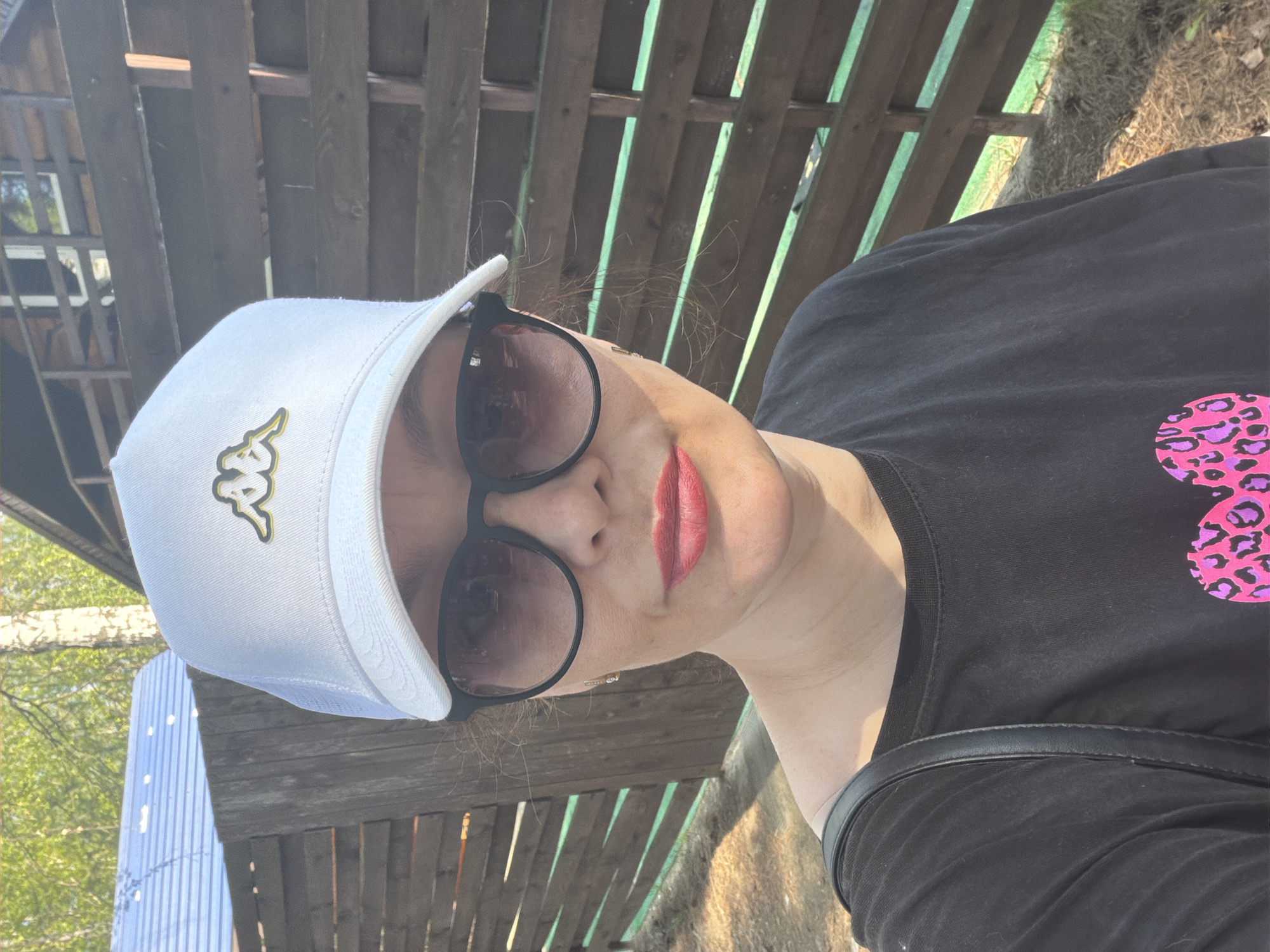 A medium shot photograph captures a woman wearing a white Kappa hat and round sunglasses. She also wears red lipstick and a black shirt adorned with a pink leopard print design. A black purse strap is visible on her shoulder. The backdrop consists of a wooden structure resembling a fence or shed. The setting appears to be outdoors with trees visible in the background. Given the query's mention of Chaikovsky, Russia, it is assumed this is the location of the photo.