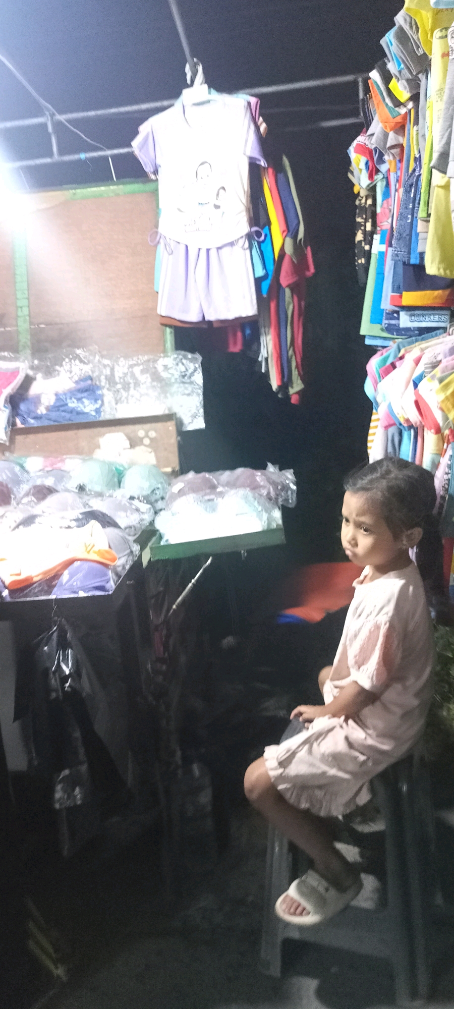 The image depicts a nighttime scene at a market stall in Bendotretek, Indonesia. A young girl, seated on a stool, is the primary human subject. She is wearing a light pink dress and white sandals. The stall displays a variety of clothing items, including children's clothes hanging on racks and folded on tables, some of which are packaged in plastic. The lighting is artificial, casting shadows and creating a somewhat dim atmosphere. The background is dark, suggesting it is outside or in an unlit area of the market.