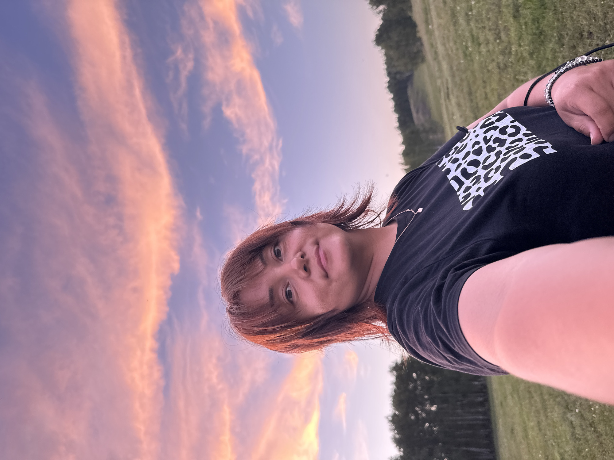 An outdoor selfie is taken by a person with reddish-brown hair and fair skin against a backdrop of a sunset sky. The person is wearing a black t-shirt with a white leopard print design on the left breast pocket area and a delicate silver necklace. The sky is filled with clouds painted in shades of pink, orange, and purple, indicative of a sunset. The background shows a grassy field with a line of trees in the distance. The lighting suggests it is either early morning or late evening.