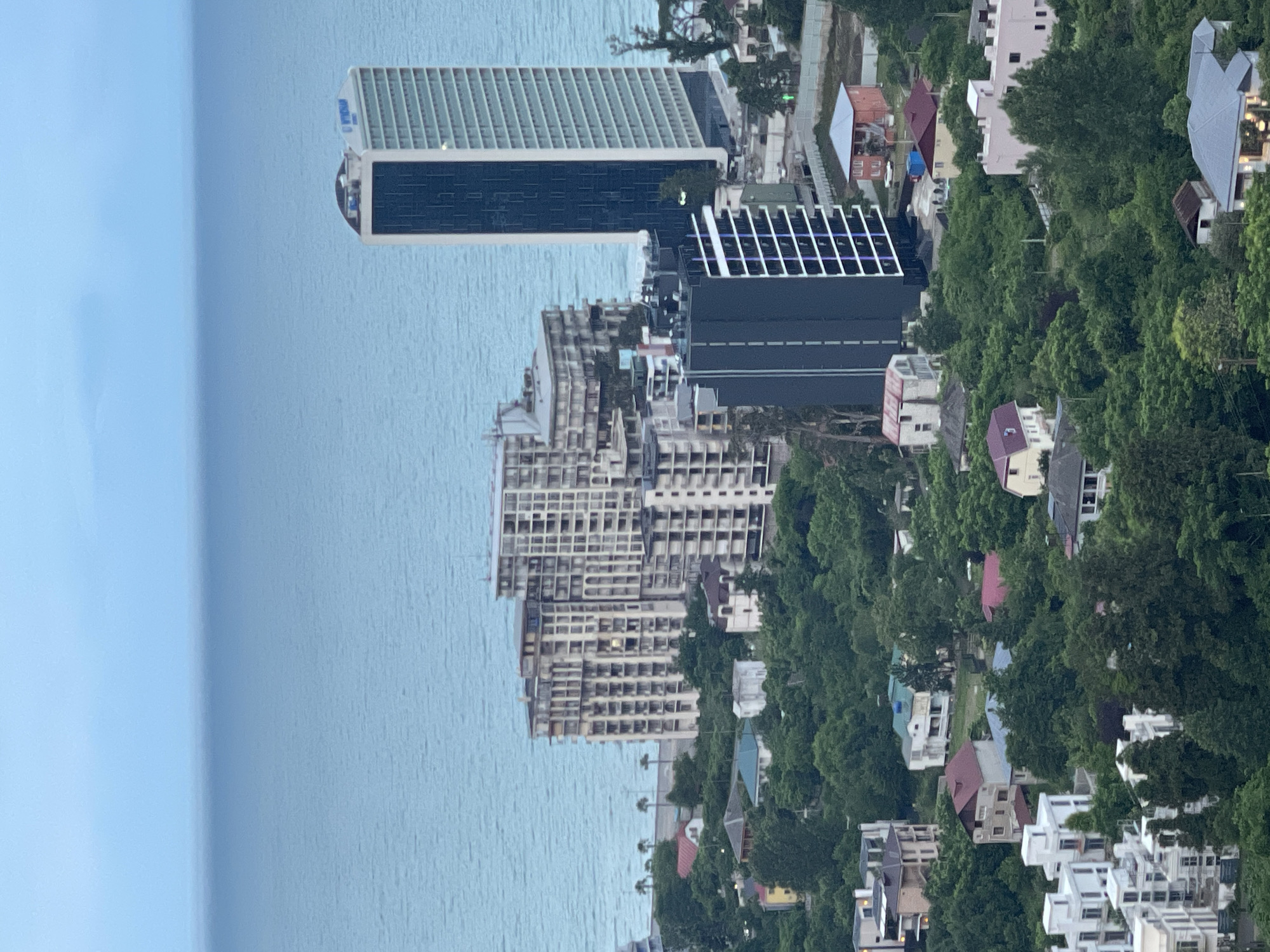 This is an aerial view of the Black Sea coast in Batumi, Georgia, featuring a mix of modern high-rise buildings and older residential structures nestled amongst dense green foliage. In the background, the calm, muted blue waters of the Black Sea stretch to the horizon under an overcast sky.
Dominating the middle ground is a cluster of apartment buildings, some appearing to be under construction or renovation, characterized by their light-colored facades and numerous balconies. To the right of these, a tall, slender, modern skyscraper with a dark blue glass facade stands prominently. In front of the larger apartment complexes, a dark grey, multi-story building with white-framed windows is visible.
The foreground and mid-ground are filled with a mosaic of houses, villas, and smaller apartment blocks. These range in style from contemporary white structures with angular designs to older, more traditional homes with red or brown tiled roofs. The buildings are interspersed with mature trees and lush green vegetation, creating a park-like setting. A few palm trees are visible along the coastline in the distance. The scene suggests a coastal urban environment with a strong presence of natural greenery. No people are discernible in the image.