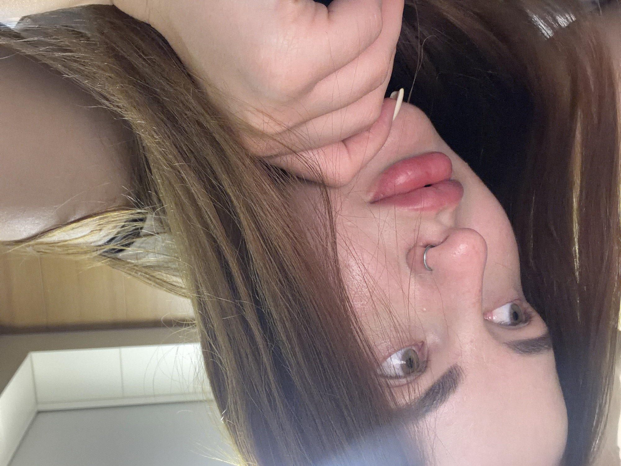 A close-up, upside-down portrait features a person, likely female, with long, light brown hair framing her face. Her head is tilted, and she is looking upwards and slightly to the right. Her eyes are light-colored, possibly hazel or light brown, with visible whites at the bottom of the iris. She has defined, dark eyebrows and a prominent nose adorned with a silver septum piercing. Her lips are full, appearing tinted pink or reddish, and are slightly parted. One hand, with light-colored fingernails, is positioned near her mouth and chin, partially obscuring that side of her face. The background is blurred but shows an indoor setting with neutral tones, suggesting a wall and ceiling. No specific details in the image indicate the location beyond being indoors. There is no visible text. The overall expression is neutral or contemplative.