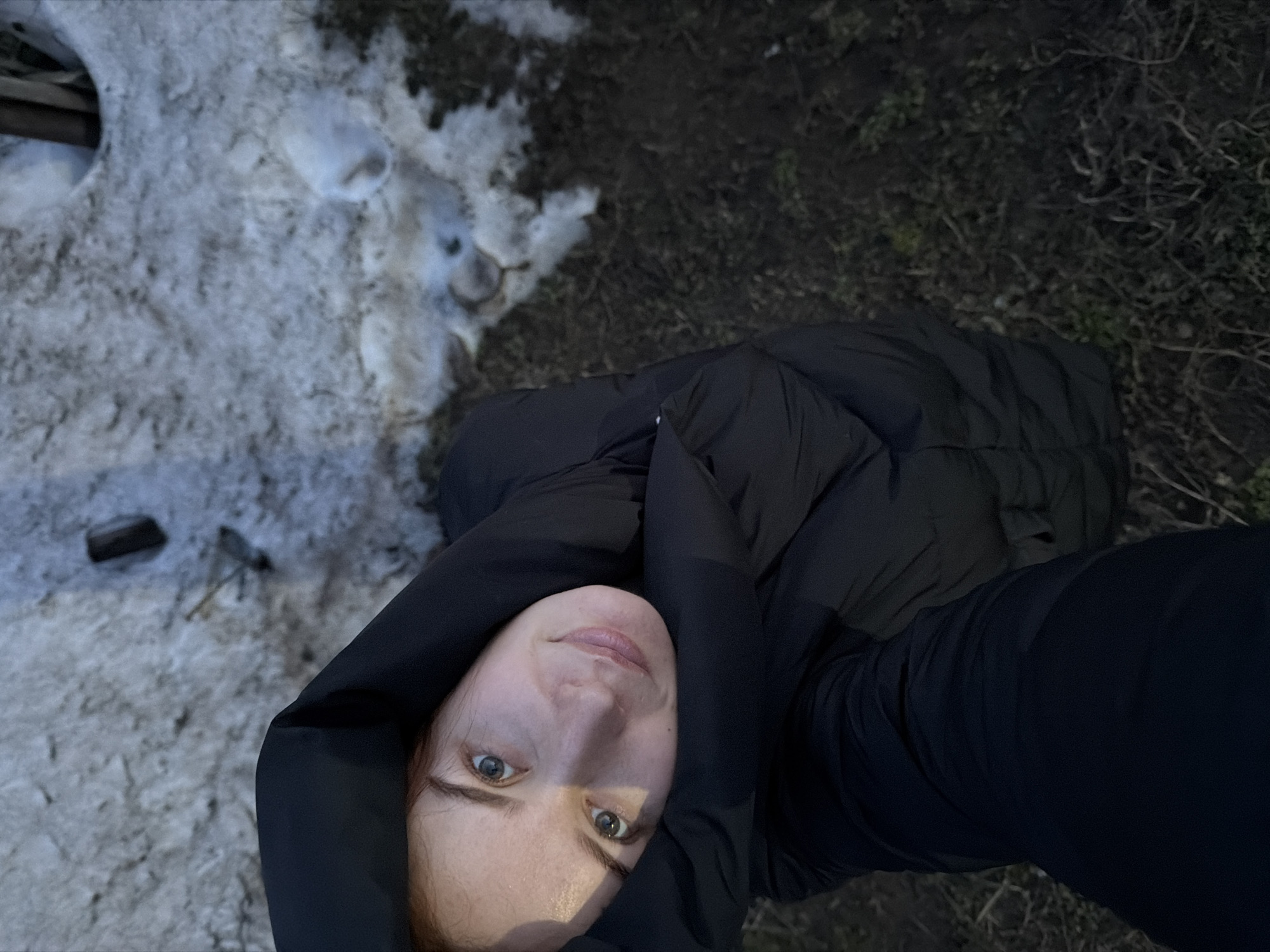 A selfie taken outdoors shows a fair-skinned person wearing a black hooded puffer jacket. To the left of the person is snow, and to the right is dark soil covered with dead grass. The location is likely Chaikovsky, Russia based on the question's request.