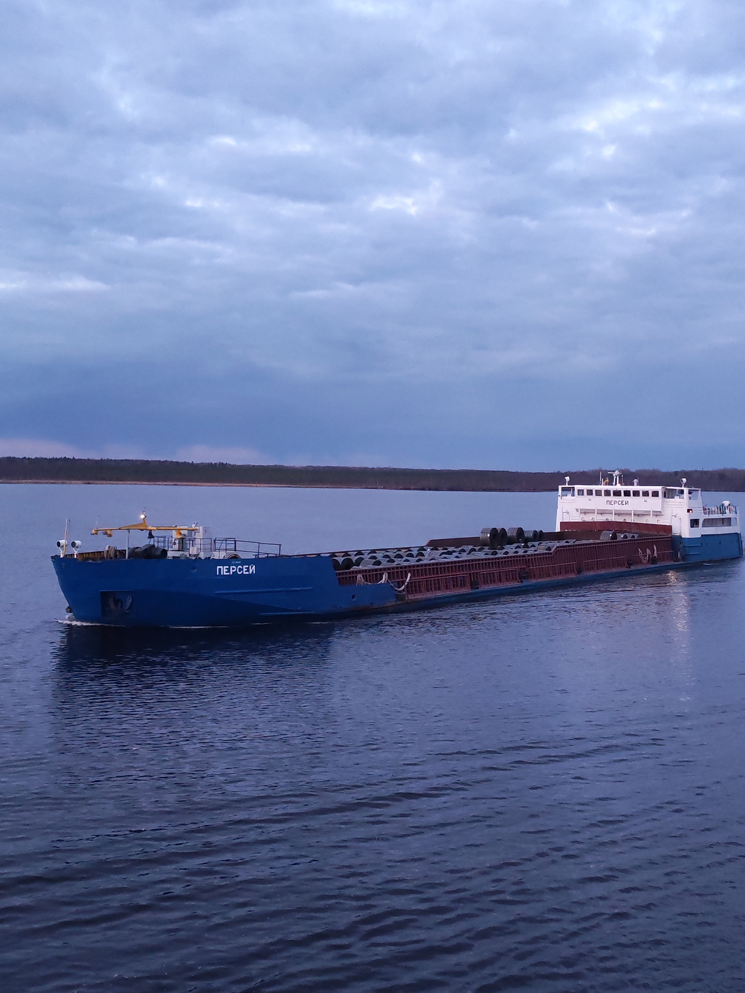 A long blue and white cargo barge, named "ПЕРСЕИ" in Cyrillic script, is sailing on a large body of water. The barge is loaded with what appears to be coils of metal material. The water is dark blue with gentle ripples. In the background, a strip of land with trees is visible under a cloudy sky. The location may be Kirgody, Russia, as specified.