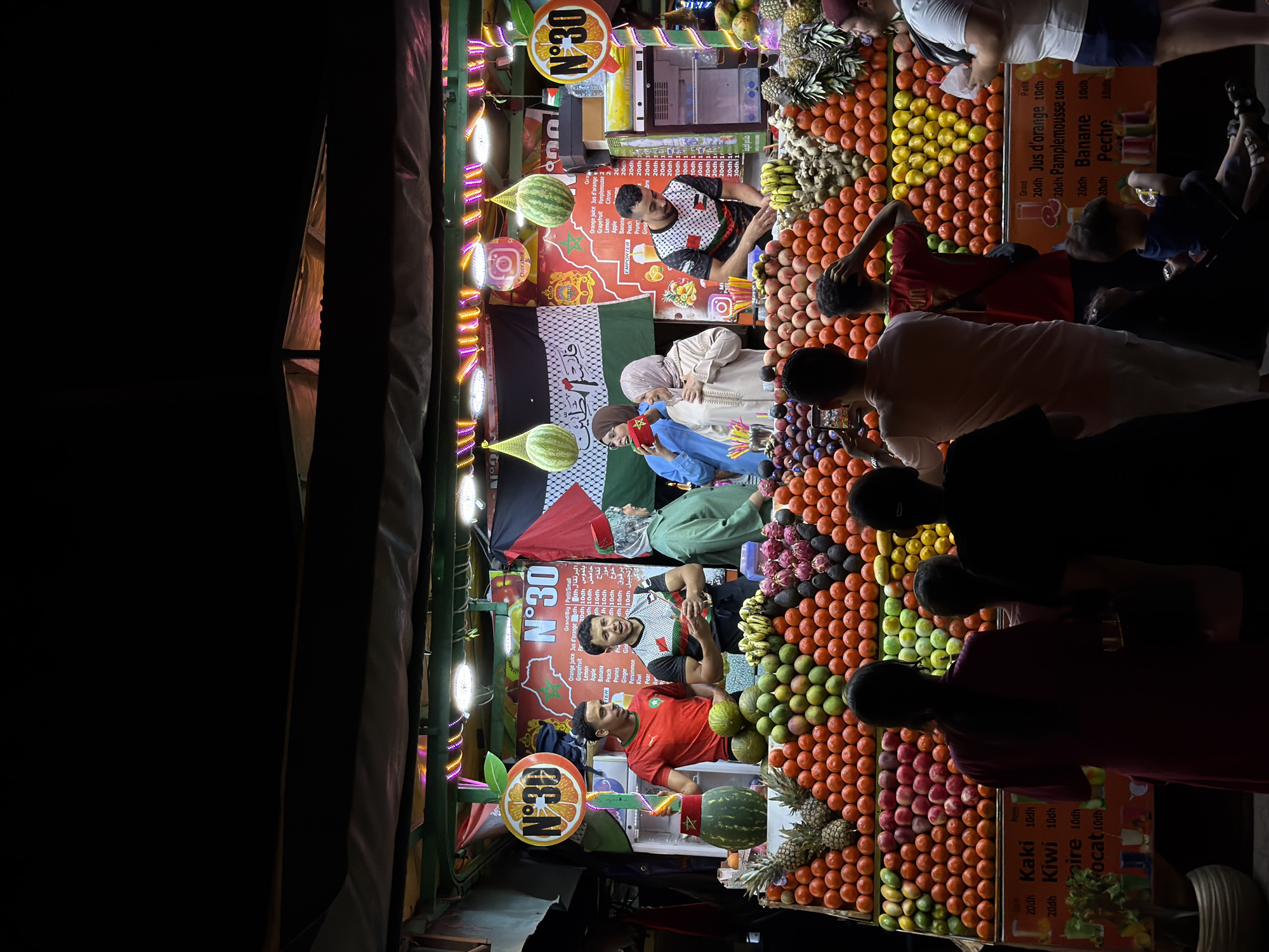 This is a nighttime photograph of a fruit juice stand in Djemaa el-Fna square, Marrakesh, Morocco. The stand is illuminated by colorful lights and displays a wide assortment of fresh fruits, including oranges, apples, bananas, pineapples, and dragon fruit. Three men are working behind the counter, preparing juices. They are wearing athletic shirts, two with a checkered pattern and one red shirt. Two women, one wearing a light-colored hijab and a traditional kaftan, and another in a light blue garment, are standing on the left side of the stand, possibly customers. A Palestinian flag and the Moroccan flag are displayed on the stand, along with signage indicating juice prices in Moroccan dirhams. The background is dark, suggesting it's late at night. Several people are visible in the foreground, observing the fruit display and the workers. A young child is sitting on the ground in the lower right portion of the image. The overall scene depicts a bustling and vibrant atmosphere typical of the square at night.