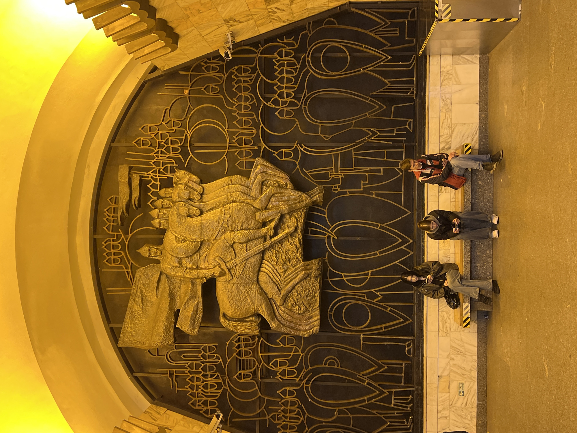 The image shows a large, ornate entrance in what is likely a subway station in Saint Petersburg, Russia. The entrance is arched and features a large, bronze relief sculpture of what appears to be a historical or mythological figure on horseback. The metalwork surrounding the sculpture consists of stylized, geometric patterns.
Three individuals are sitting on a bench against the right side of the entrance. The first is wearing a jacket, jeans, and shoes. The second individual is wearing a black jacket and jeans. The third is wearing a jacket and grey pants. The environment is well-lit with a yellow-toned light. The floor is a combination of marble and light-colored concrete. A surveillance camera is mounted on the wall above the opening.