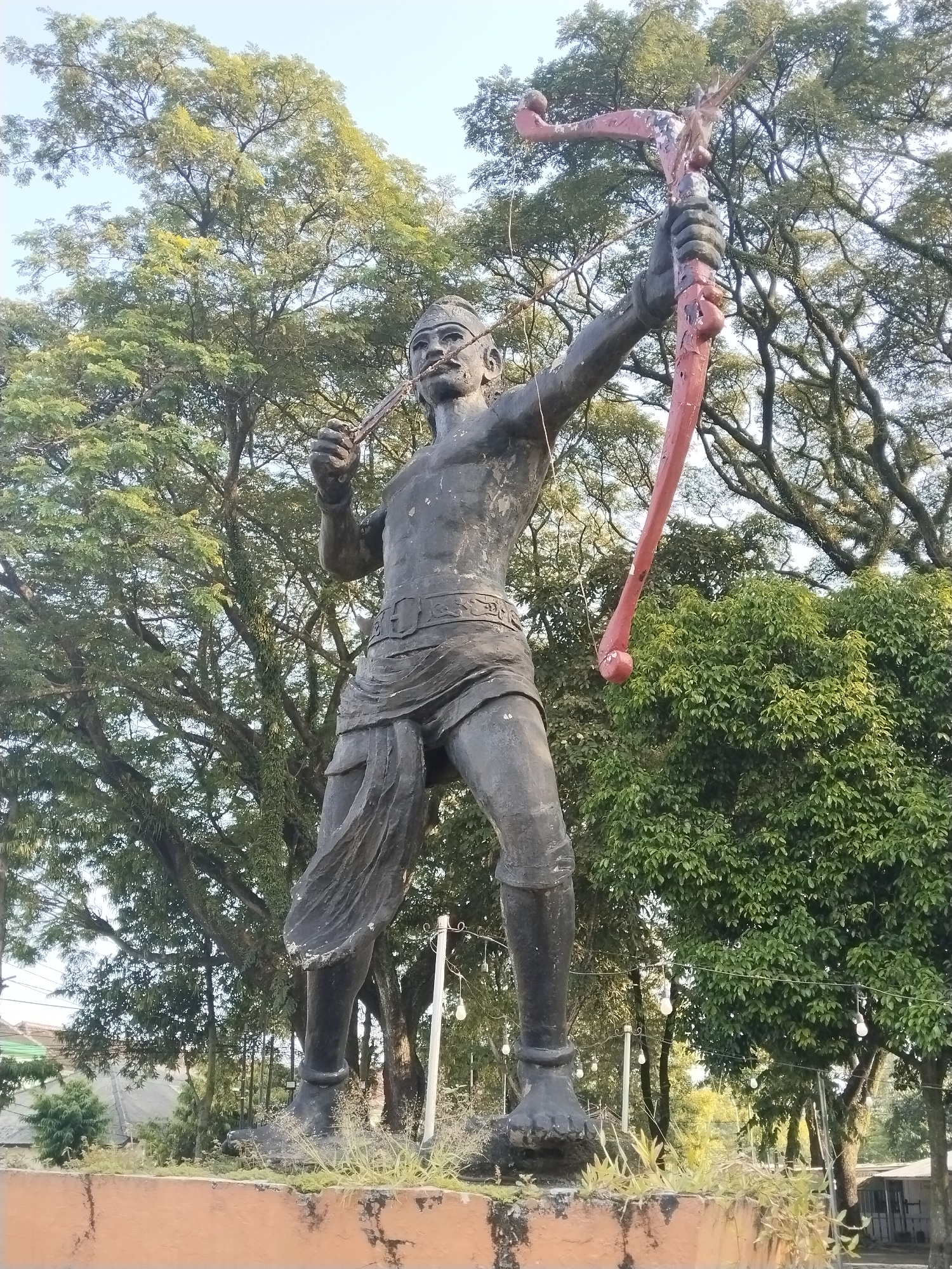 The image displays a large, weathered statue of a male archer, likely a warrior or legendary figure, set outdoors in what appears to be a park or public square. The statue, predominantly dark gray, stands dynamically, depicting the moment an arrow is drawn back in a red-painted bow, poised for release. The archer holds the bow with his left hand and the arrow with his right, featuring a determined facial expression, complete with a mustache and a simple headdress. He wears traditional attire, including a cloth draped around his waist.
The statue rests on a raised, rough, light-orange and brown base, where sparse grass and weeds have grown. Surrounding the statue and forming the background are numerous large trees with abundant green foliage and visible branches, suggesting a natural and established outdoor environment. The sky above is a clear, light blue, indicating bright daylight conditions, possibly late morning or early afternoon. In the background, particularly on the right side, string lights are visible hanging between trees, suggesting the area is illuminated at night. Hints of distant structures or buildings with rooftops can be seen through the trees, placing the monument within a populated area in Sidoharjo, Indonesia. There is no visible text in the image.
