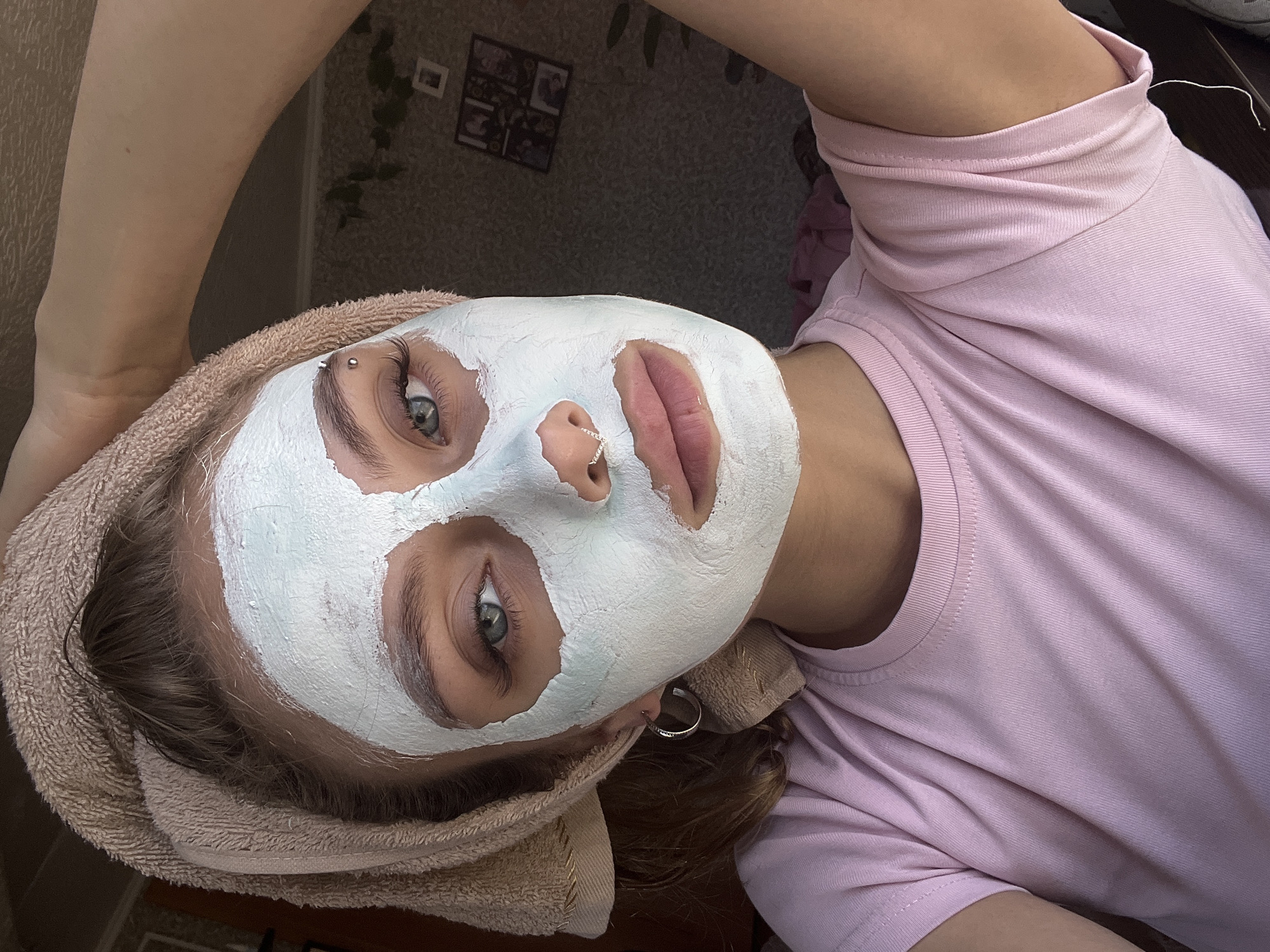 A young woman with a face mask on, a towel wrapped around her head and a pink t-shirt. The woman has blue eyes, dark eyebrows and eyelashes. She is wearing a silver hoop earring in her left ear and a nose ring. The background appears to be a room in a house. There are framed pictures on the wall and some plants. The scene is well-lit, with natural light coming from a window. The woman is looking directly at the camera, with her right hand touching her head. The overall impression is a casual selfie taken at home. The location of Фоки, Russia is not ascertainable from the image.