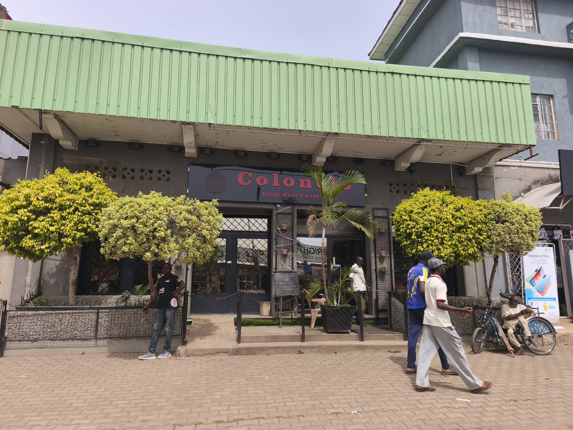 This full shot captures a street view of "Colony Restaurant & Bakery" in Jos, Nigeria, during clear daylight hours. The establishment is marked by a prominent green corrugated metal overhang above a dark grey facade. A black sign with red lettering clearly displays "Colony Restaurant & Bakery."
Several men are visible in the foreground and around the entrance. On the far left, a man in a black t-shirt and jeans stands leaning against a low metal fence, which encloses decorative, lush green and yellow-leafed bushes. In the center, near the entrance, an A-frame chalkboard menu lists items such as "CHICKEN WING," "FOUR SEASONS PIZZA," "CHICKEN BURGER," and "CHIPS." Potted plants, including a palm, adorn the entrance area.
Two men are walking in the right foreground; one wears a blue and yellow top, and the other is in a light-colored t-shirt and grey sweatpants. Further to the right, a man with a beard sits in a hand-pedaled tricycle, next to a blue bicycle, seemingly waiting or resting. A bright promotional poster for "REDMI A7 Pro" is displayed on the wall behind him. The ground in the foreground is paved with interlocking bricks, typical of a busy urban street. The overall impression is a lively commercial area.