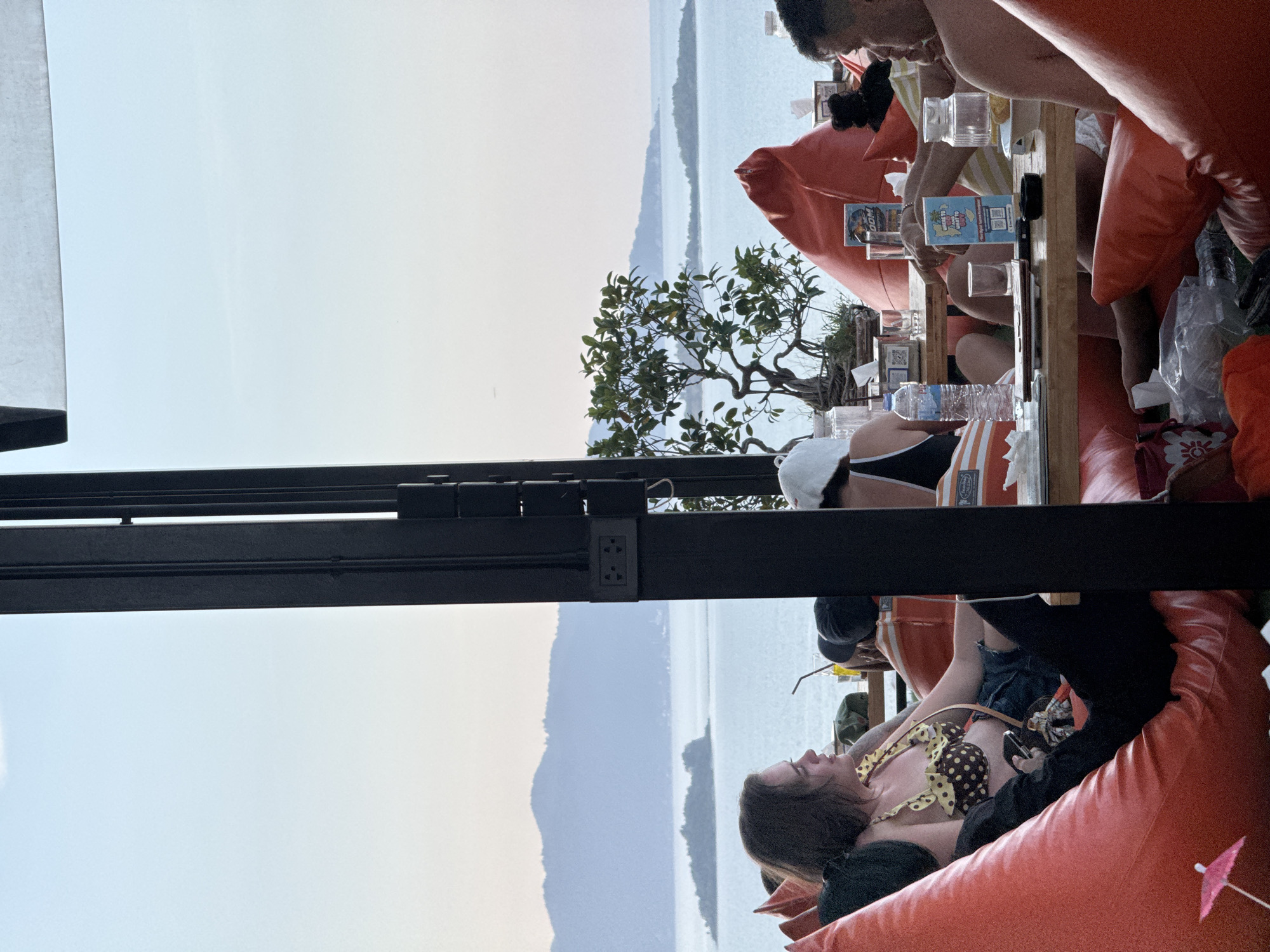 This image depicts a group of people seated at a table in an outdoor dining area overlooking a body of water with islands and mountains in the distance. The setting appears to be a restaurant or bar in a tropical location, likely Ko Samui District, Thailand, given the style of the foliage and the distant landscape. The people are casually dressed, with some wearing swimwear. They are seated on large, orange beanbag chairs and at a wooden table laden with drinks and menus. The lighting suggests it is late afternoon or early evening. The overall atmosphere is relaxed and social, consistent with a vacation or leisure setting.