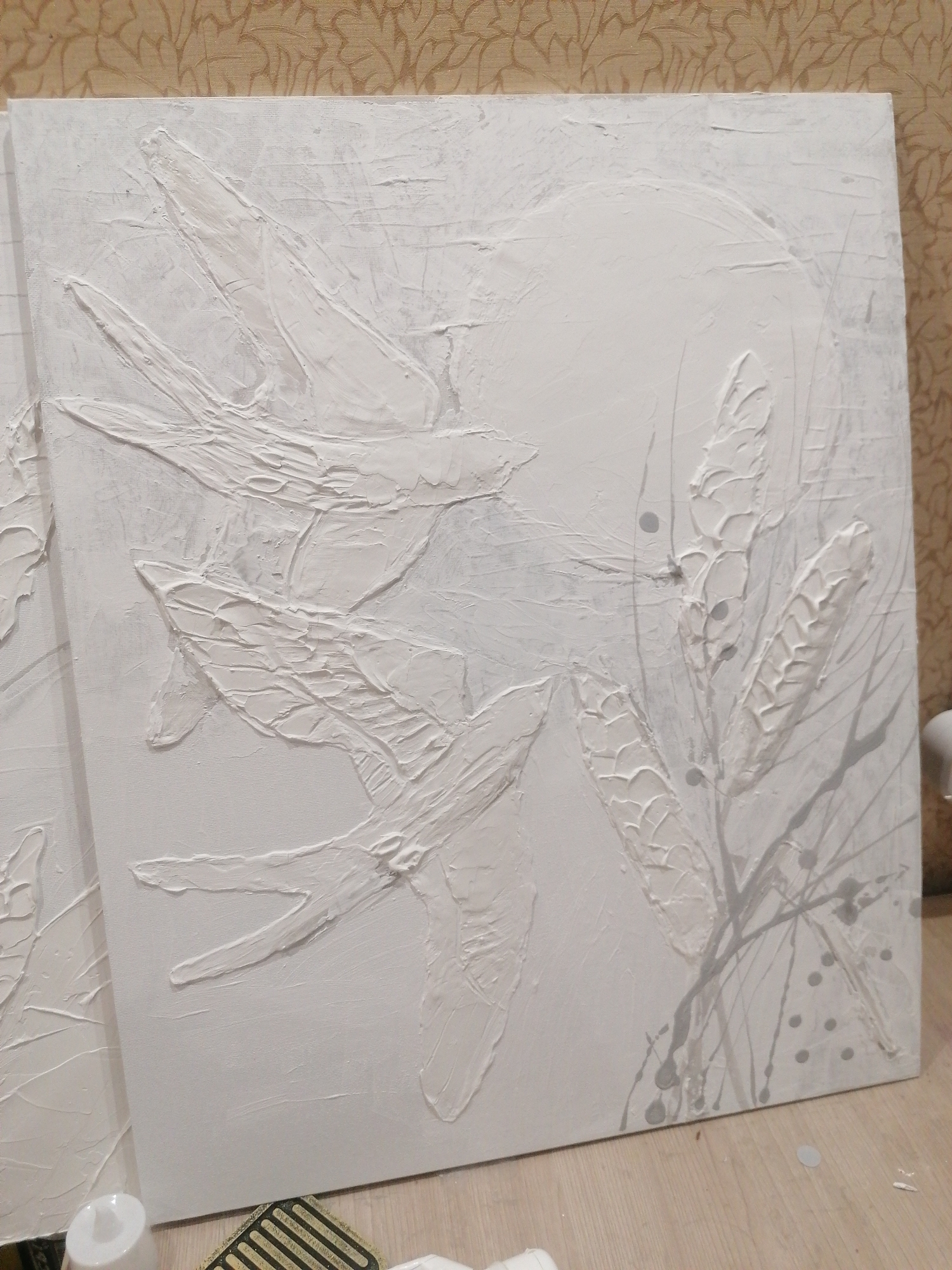 The image shows a textured, three-dimensional artwork on a canvas, primarily in white with subtle gray accents. The artwork depicts several stylized birds in flight, with their wings outstretched and rendered with thick impasto. There are also elements resembling plant stalks or seed heads, also in textured white, positioned on the right side of the canvas. Gray paint has been splattered and dripped onto the canvas, particularly around the plant elements and forming scattered dots. The canvas is leaning against a wall with a textured wallpaper featuring a repeating floral or leaf pattern. The lower portion of the image shows a wooden floor. In the foreground, a white cylindrical object, possibly a can of paint or a tool, is visible. Another object with a dark grid pattern is partially visible. There are no people or identifiable landmarks of Angarsk, Russia, present in the image.
