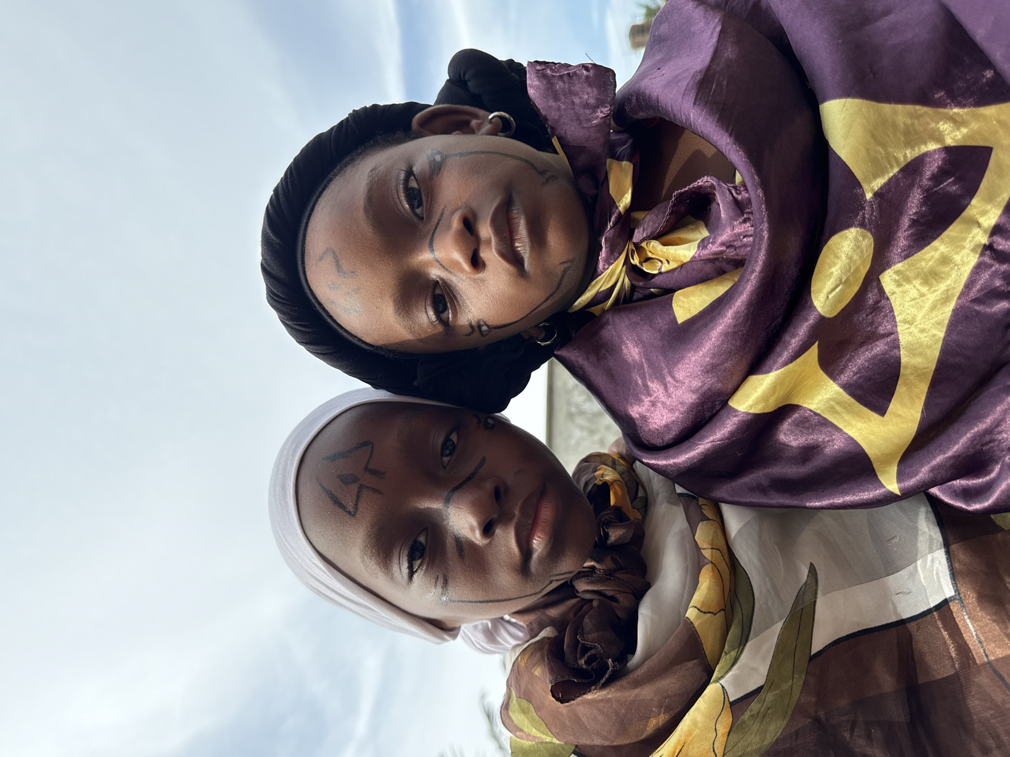 Two children, appearing to be of similar age and ethnic background, are pictured in a close-up, waist-up shot against an overcast sky. Both children have intricate black face paint, including lines on their foreheads, cheeks, and around their eyes. They are wearing head coverings and draped with patterned fabrics around their necks and shoulders. The child on the left wears a white head covering and a brown and gold patterned fabric. The child on the right wears a black head covering and a shimmering purple and gold patterned fabric. The background is a plain, pale sky with some visible clouds. There are no discernible urban structures or elements specific to Ado, Nigeria, visible in the frame.