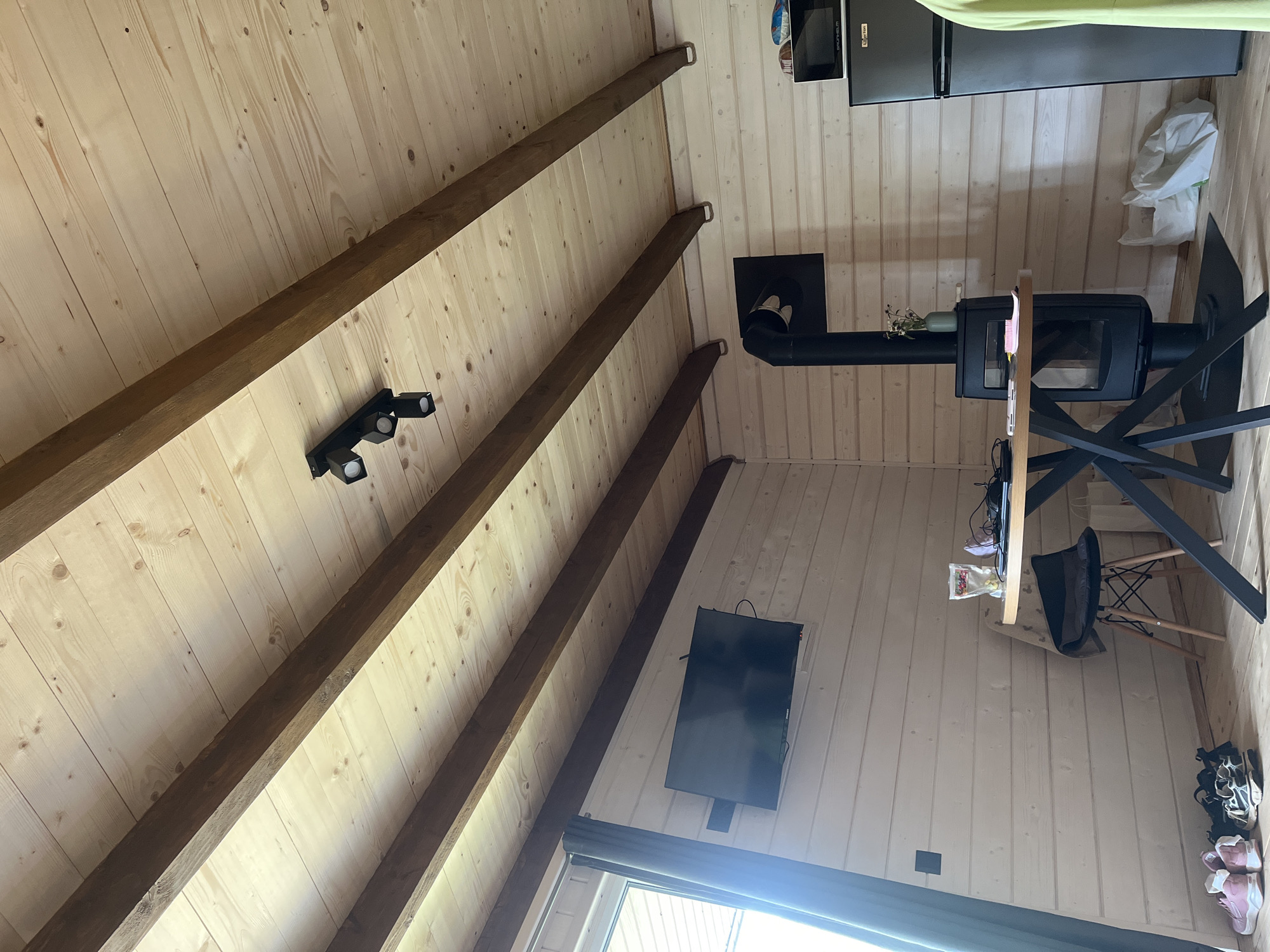 The image shows the interior of a room with wooden paneled walls and ceiling. Exposed wooden beams run across the ceiling. A black track lighting fixture with three adjustable spotlights is mounted on the ceiling. On the wall, a flat-screen television is mounted. A modern wood-burning stove with a black pipe is visible in the corner. A small round table with a black metal base is set up with a chair. On the table, there are some items including a laptop, a bag of snacks, and a small vase with flowers. A refrigerator is situated next to the stove. On the floor near the wall, there are several pairs of shoes. The room appears to be a living space or a common area. Based on the general style and common amenities found in such locations, this could be a room within a guesthouse or a private residence in a rural or semi-rural area. Given the information that the location is in Mykulychyn, Ukraine, this scene likely depicts a room in a Carpathian mountain region guesthouse, often catering to tourists.