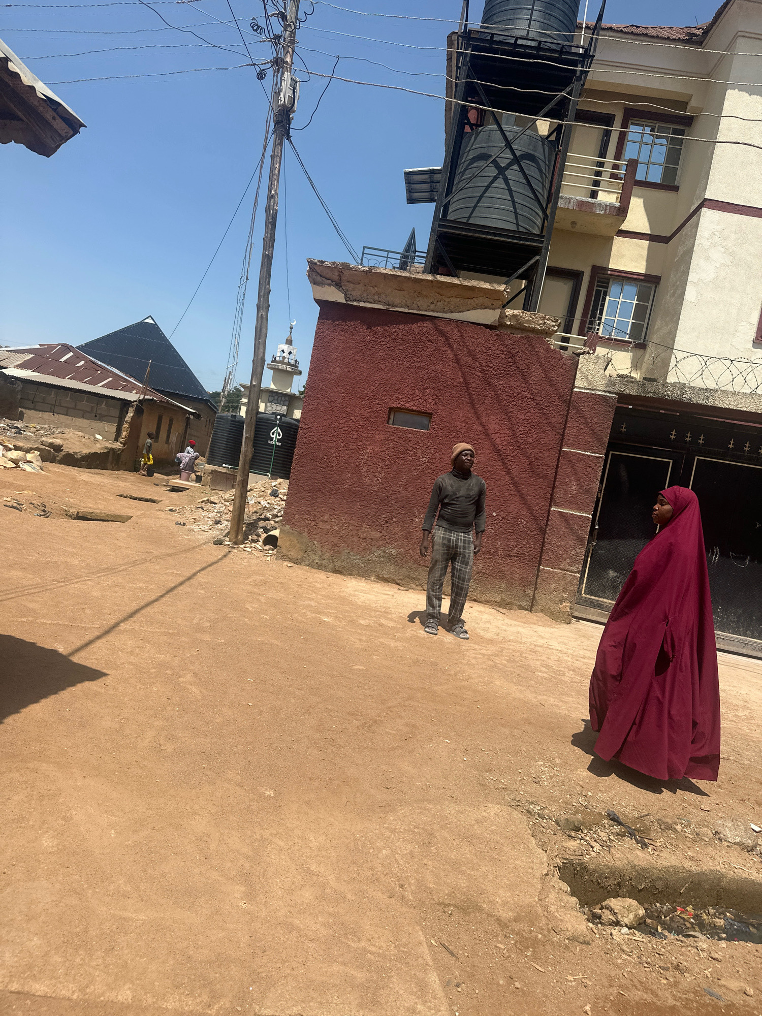 In Barakin Lerawi, Nigeria, an outdoor scene unfolds on a dirt road under a clear blue sky. A dark-skinned man wearing a light brown beanie, a grey long-sleeved shirt, and checkered pants stands on the right side of the frame. He is facing left, looking upwards. To his right and slightly in front, a person draped in a maroon hijab and abaya stands, also facing left and looking upwards.
Behind them, a multi-story building with cream-colored walls and brown trim features multiple windows, some with reflective glass. A metal structure supporting two large black water tanks is attached to the side of this building. To the left of the building, a red-walled structure with a small rectangular window is visible. Further to the left, several single-story buildings with corrugated metal roofs are present. A mosque with a white minaret and a crescent moon on top is visible in the background between the buildings. A utility pole with numerous wires stretches across the upper portion of the image. The ground is predominantly composed of reddish-brown dirt with scattered rocks and debris. Shadows are cast on the ground, indicating a sunny day.