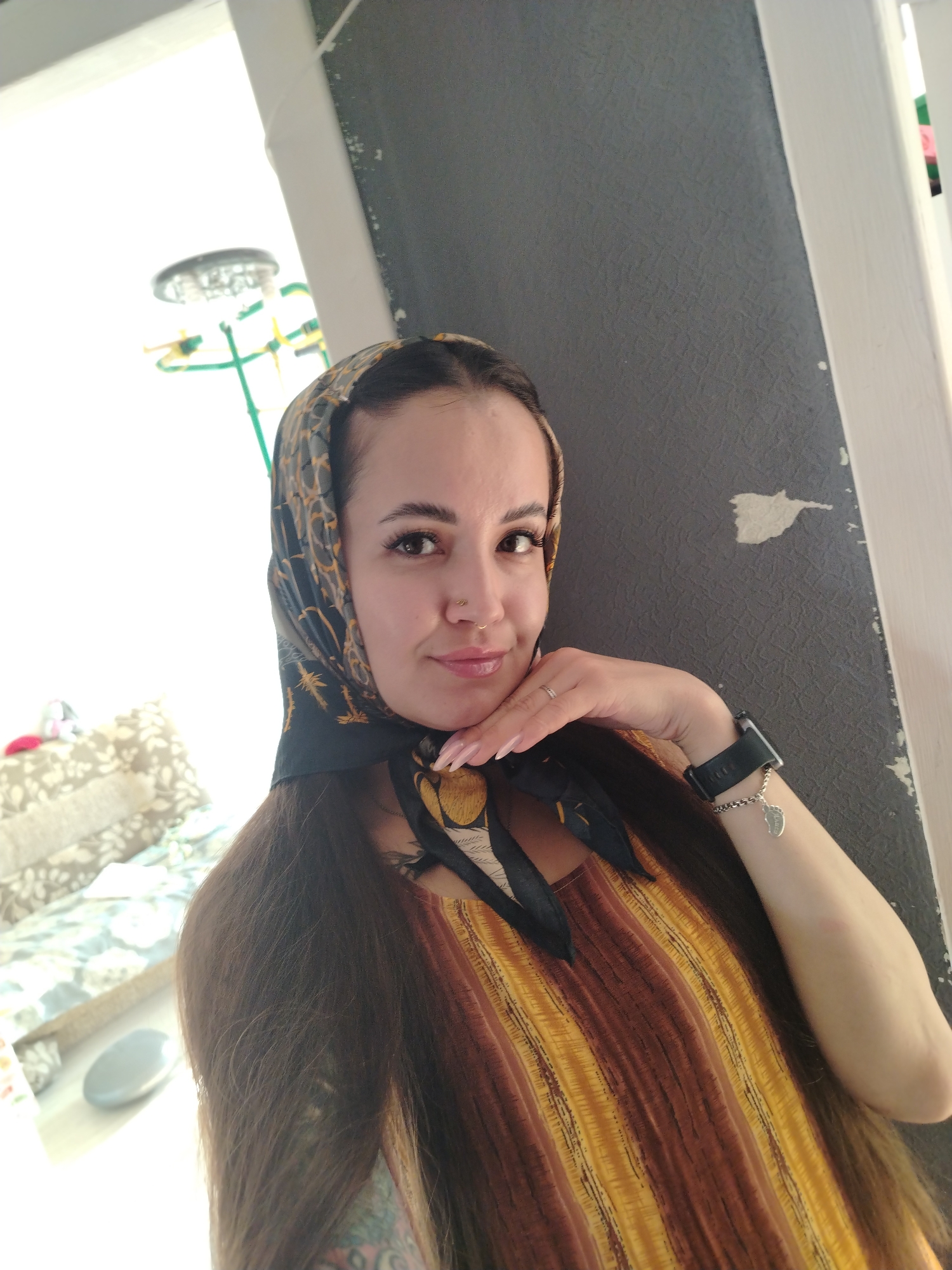 In Noviy, Russia, a woman with long, dark hair tied in a headscarf is pictured indoors, leaning against a grey, textured wall. She is wearing a striped brown and yellow dress and has a smartwatch on her left wrist with a silver charm. Her right hand is resting on her chin, and she is looking at the camera with a slight smile. The background shows a brightly lit area with what appears to be furniture and a rack with green and yellow elements.