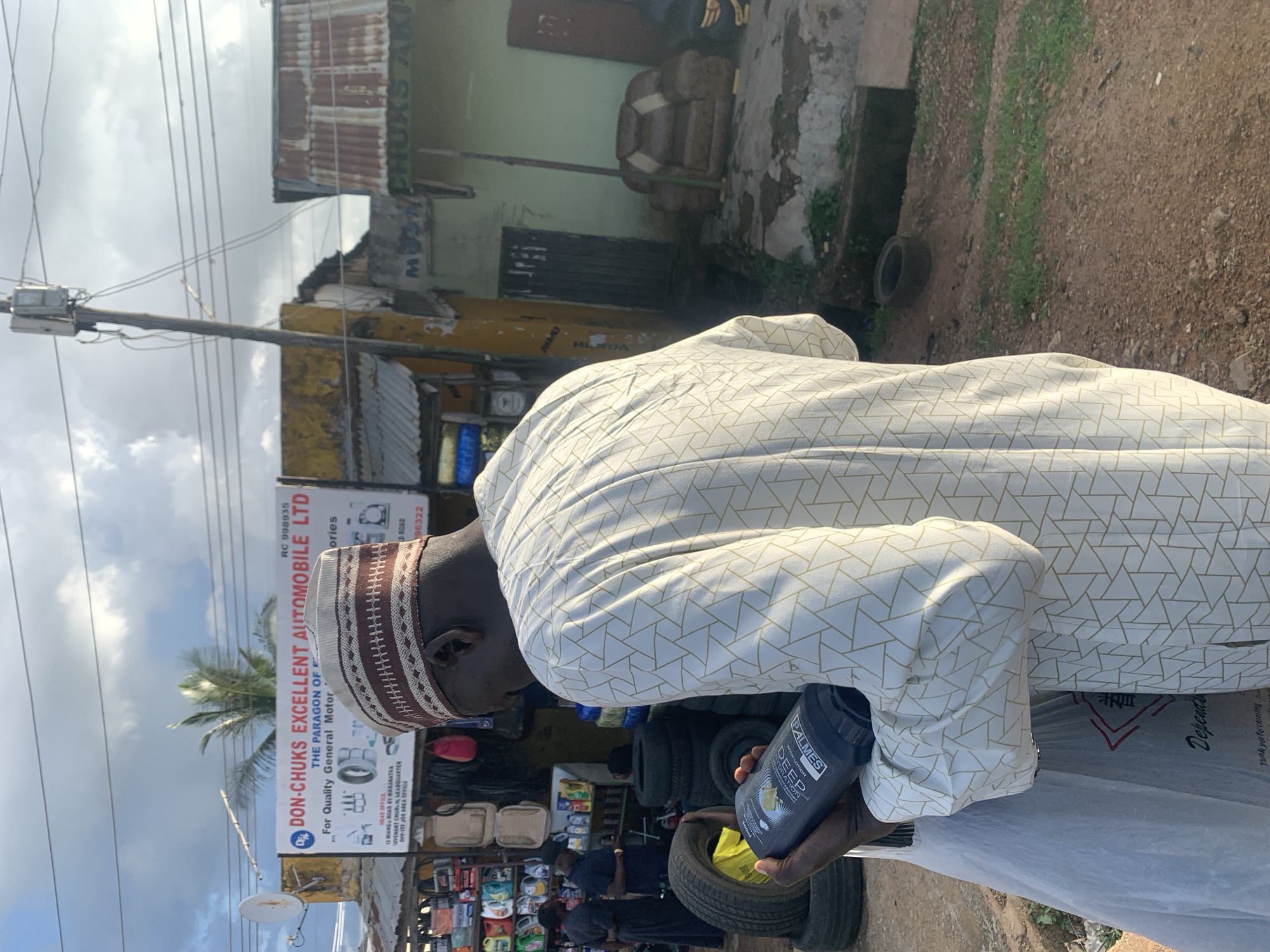 The image depicts a scene in Jos, Nigeria, with a person in the foreground wearing a white and gold patterned robe and a decorative cap. The person is holding a dark blue bottle of Palmes Body Lotion. The background shows a street scene with a shop. A sign for "DON-CHUKS EXCELLENT AUTOMOBILE LTD" is visible, indicating it's a business selling automotive parts. Other signs and shelves filled with various products are also present, suggesting a busy marketplace or roadside stall. Tires are stacked in the background, and there's a general sense of an outdoor commercial area. The lighting suggests it's daytime, possibly late morning or afternoon, with a mix of sunlight and clouds in the sky. The overall atmosphere appears to be that of a typical commercial street in a Nigerian city.