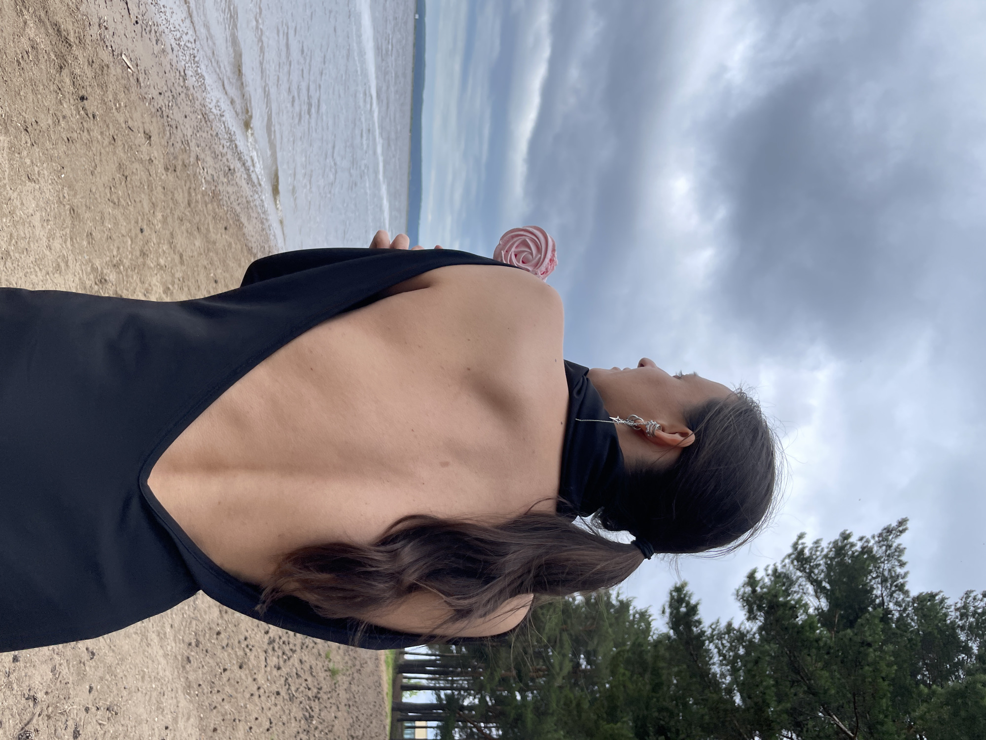 A woman with her back to the camera stands on a sandy beach, holding a pink lollipop. She is wearing a black dress with a deep V-neckline exposing her back. The ocean is to her left, and a forest of pine trees is to her right. The sky is cloudy. The location is the Kama River embankment in Chaikovsky, Russia.