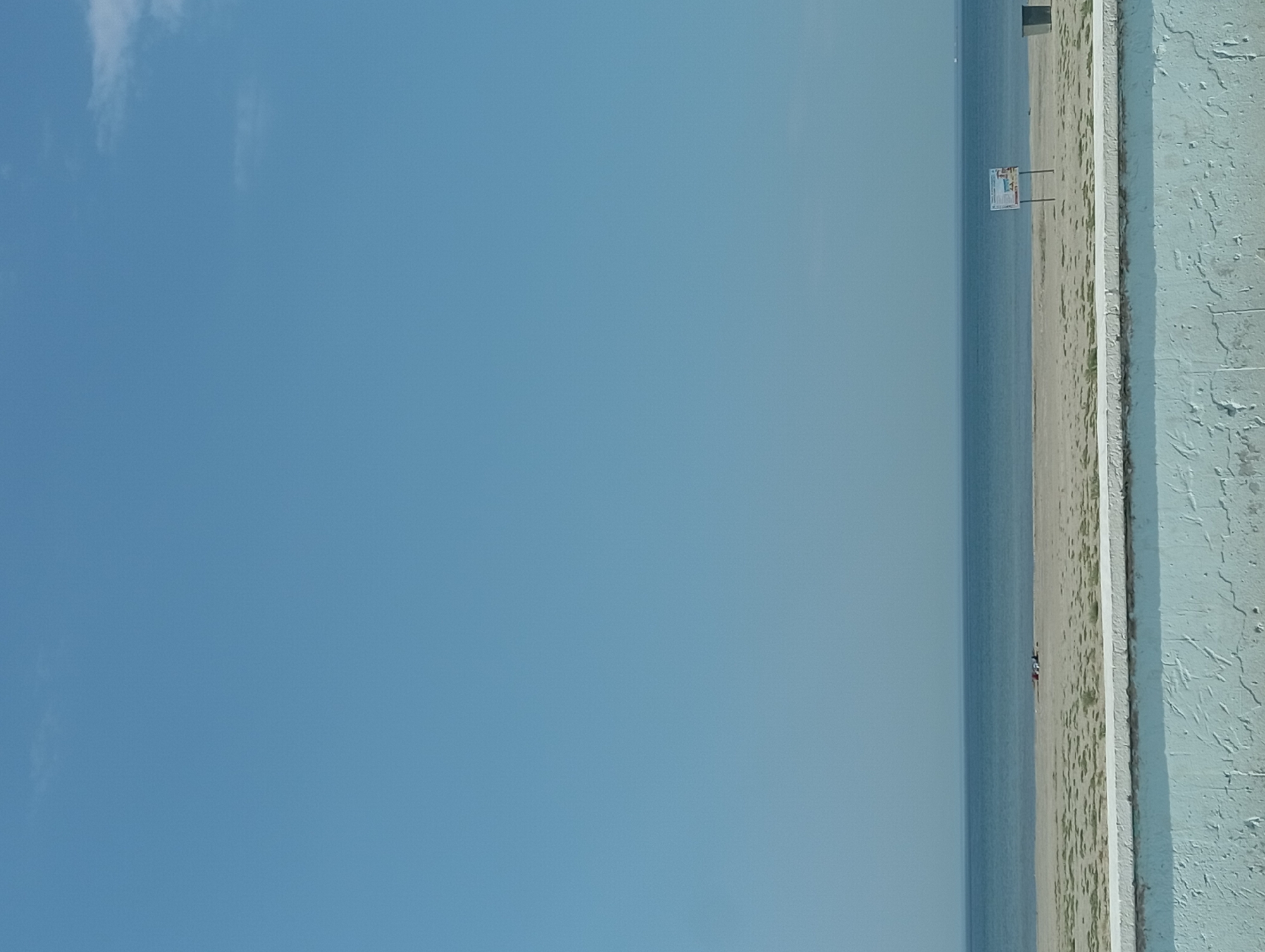 This is a photograph taken from a low vantage point, looking out over a beach towards the sea. In the foreground, a textured, light blue concrete wall runs horizontally across the bottom of the frame. Beyond the wall is a sandy beach with sparse green vegetation. In the middle ground, there is a calm body of water, likely the Caspian Sea, stretching to the horizon. The sky is clear and pale blue, with a few wisps of clouds near the top right.
On the beach, there are a few indistinct figures, possibly people sitting or lying down. Further to the right, there is a rectangular signpost with a white sign that appears to have text and images on it. To the far right, there is a dark gray trash receptacle. The scene suggests a quiet day at the beach. The location is the city of Ömirzaq, Kazakhstan.