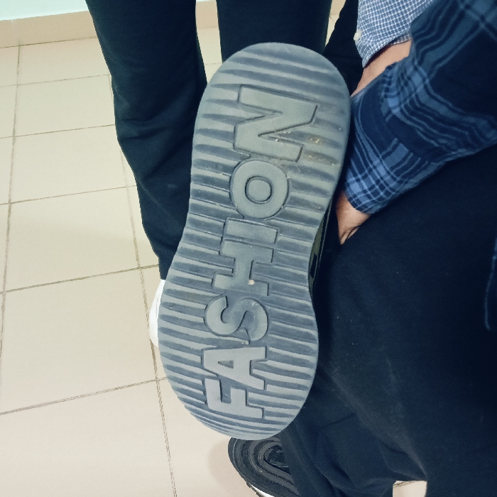 An indoor scene is captured from a top-down perspective, focusing on footwear and lower extremities. The prominent subject is the upturned dark gray sole of an athletic shoe, likely a sneaker, positioned centrally and oriented vertically. The word "FASHION" is embossed in large capital letters on the sole, accompanied by horizontal ridged patterns. The shoe is situated amongst the legs of at least two individuals. One individual wears black pants and a blue plaid shirt, with a hand visible near the shoe. Another individual's leg, clad in black pants, is visible on the left. A portion of a black shoe with a white sole is visible at the bottom. The floor consists of light-colored square tiles, and a light blue-green wall or baseboard is present in the upper left background. No specific identifying features are present to determine a precise location within the city of Chaikovsky, Russia.