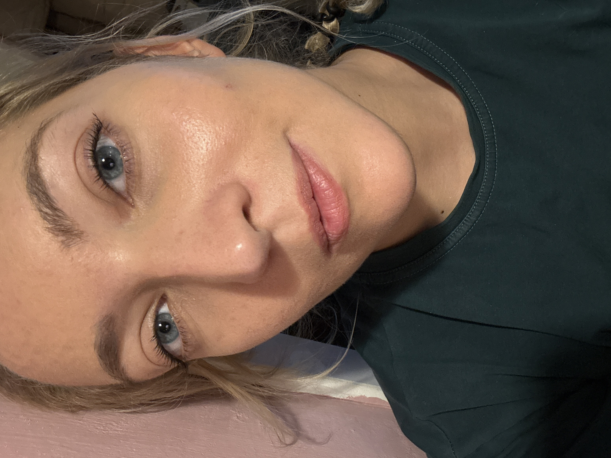 A close-up, sideways portrait of a young Caucasian woman with fair skin, blonde hair, and blue eyes. She is wearing a dark green t-shirt. The woman's face is angled towards the camera, and her lips are slightly parted. Her eyes are open and looking directly at the viewer. Her eyelashes are dark and appear to be enhanced with mascara. The background is blurred, but hints of a pink surface and darker fabric suggest an indoor setting. The lighting is soft, creating a subtle glow on her skin, and the overall mood is relaxed and intimate. There are no visible landmarks or text in the image. The location context is Minsk, Belarus.