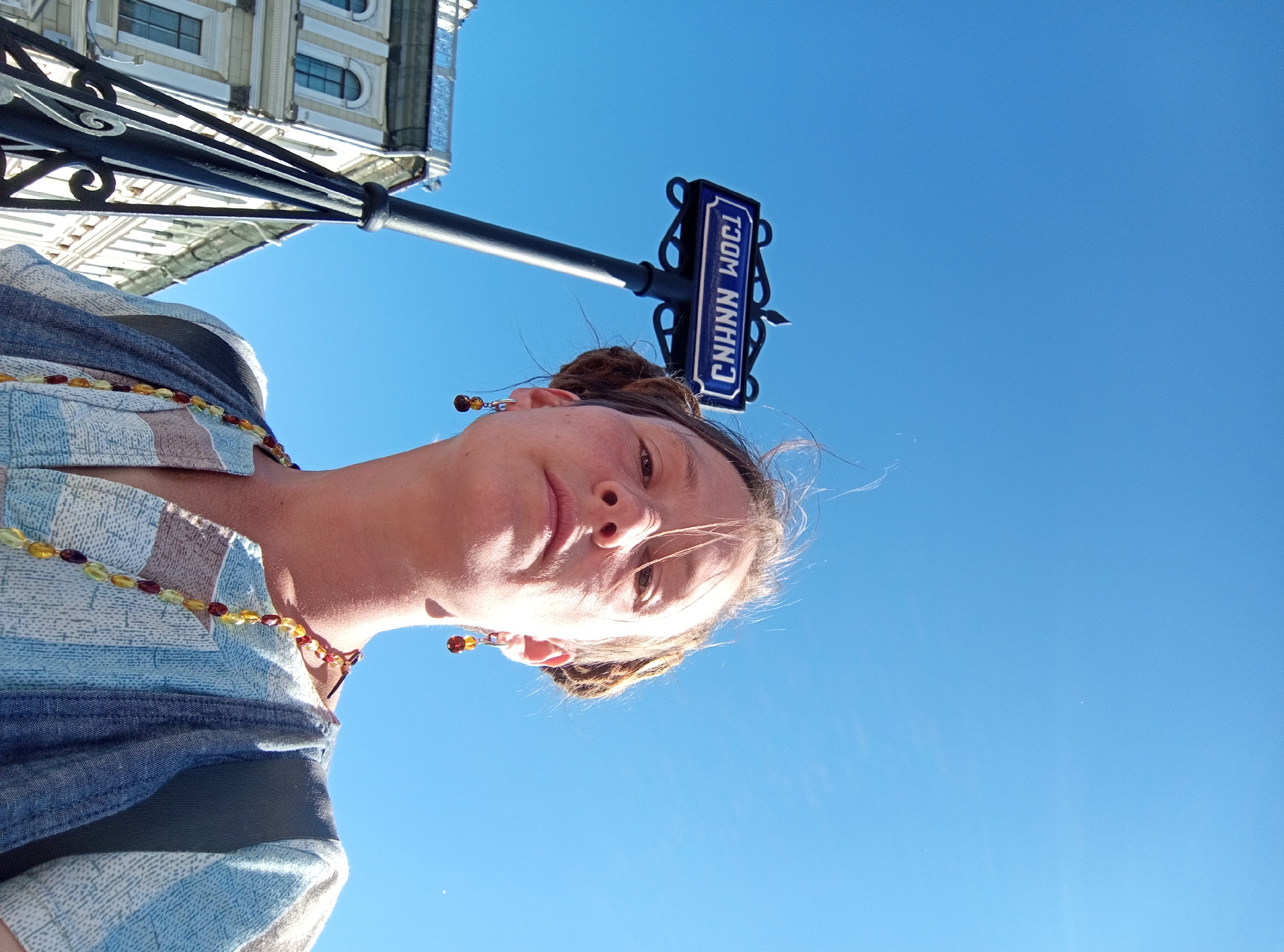 The image shows a low-angle, close-up selfie of a young person against a bright blue sky. The person is wearing a light-colored, patterned shirt and a beaded necklace. They have earrings and their hair is pulled up. In the background, a building with windows and decorative architectural elements is visible, along with a street sign attached to a black lamppost. The street sign is blue with white text.
The time of day appears to be daytime, given the bright sky and sunlight casting shadows. The weather seems clear and sunny. The presence of the street sign, building, and lamppost suggests an urban setting. The visible text on the street sign reads "CHNN WOCL". This, combined with the general architectural style of the building and the style of the street sign, is consistent with a location in Saint Petersburg, Russia. The person's expression is relaxed and casual, and the overall mood is bright and outdoorsy.