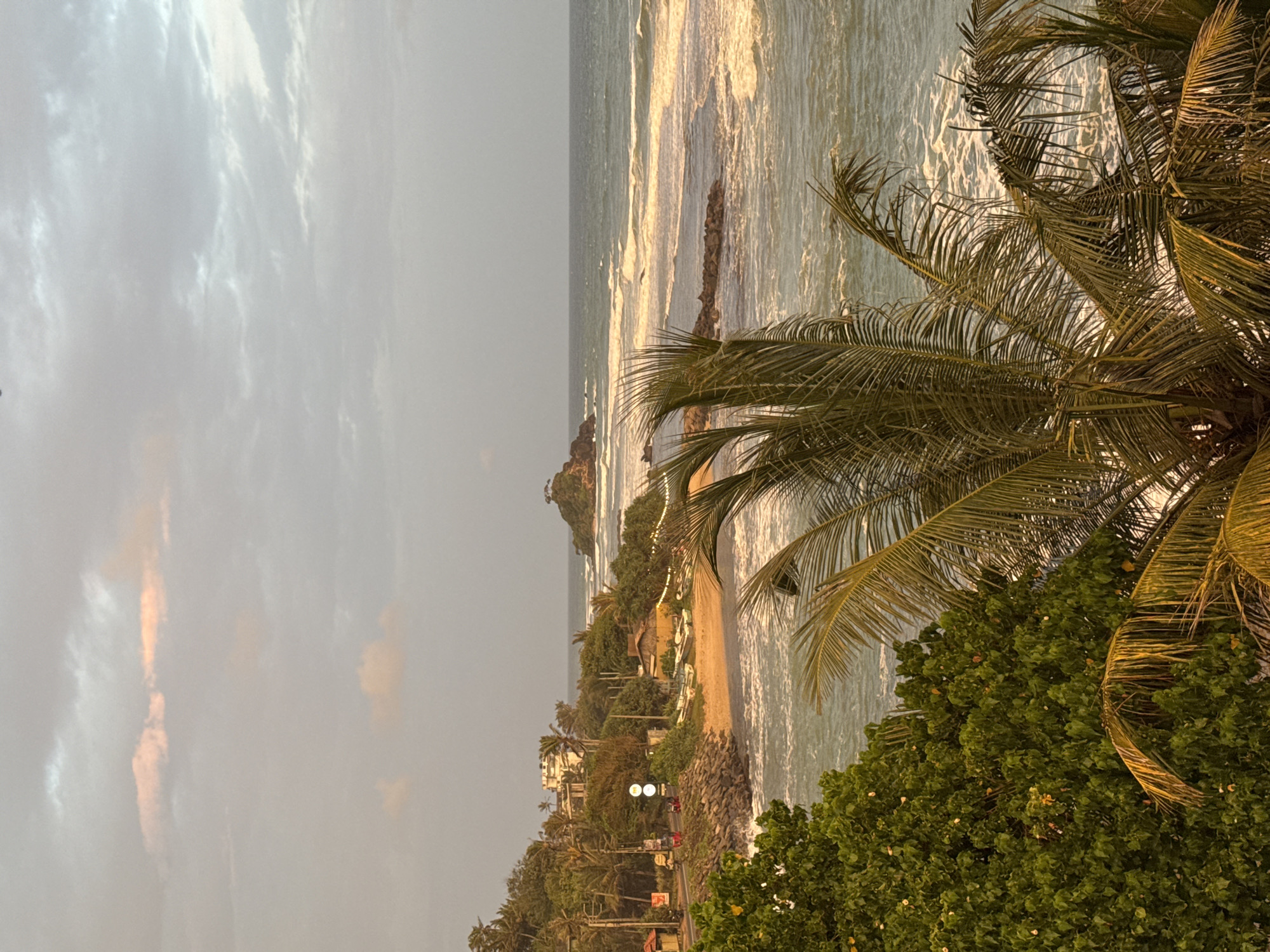 The image depicts a scenic coastal view in Ahangama, Sri Lanka. In the foreground, palm fronds from two palm trees partially obscure the view, with one tree's fronds in focus and golden-hued, while the other is slightly out of focus and greener. Beyond the trees, a sandy beach stretches along the coast, dotted with several small boats pulled ashore. A low, sandy-colored building with a thatched roof is situated on the beach, and a small cluster of lights are strung along its front.
A road runs along the edge of the beach, with a few vehicles visible, including what appears to be a red tuk-tuk. A traffic signal with two round lights is also present. Lush green vegetation, including more palm trees, covers the hillside that rises from the road and beach. In the distance, the ocean is visible, with moderate waves breaking and creating white foam. A small rocky outcrop with some vegetation and a single tree rises from the sea a short distance from the shore. The sky above is overcast with a few wisps of pink and grey clouds, suggesting either sunrise or sunset. The overall scene conveys a tropical, tranquil atmosphere.
