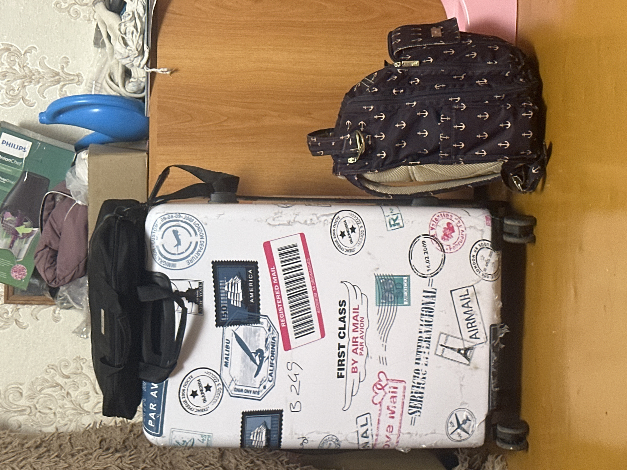 The image shows a white rolling suitcase with numerous travel-themed stickers, including postage stamps and city names like "MALIBU," "CALIFORNIA," "NEW YORK," and "LONDON." A black duffel bag is placed on top of the suitcase. To the right of the suitcase, a dark blue backpack with a pattern of anchors is leaning against a wooden surface. In the background, a blue plastic watering can and other items are visible on a shelf. The scene appears to be an indoor domestic setting, possibly a room in a house or apartment in Загородный, Russia. The flooring is a textured yellow. There are no people present.